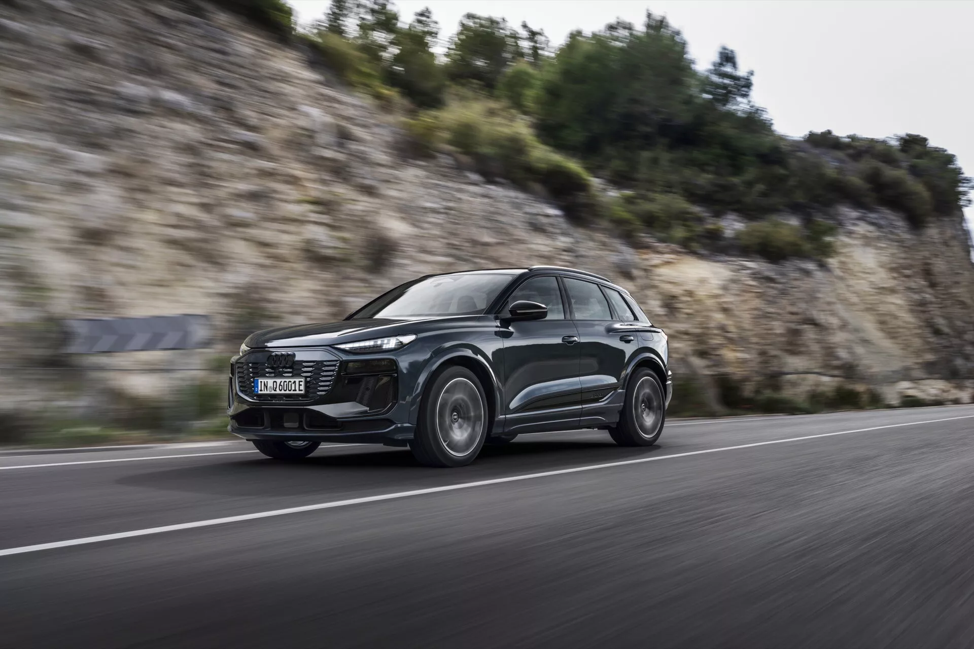Foto dinâmico do Audi Q6 e-tron 2024 em movimento de um ângulo baixo e frontal, mostrando seu design SUV aerodinâmico elegante, faróis LED modernos e rodas de liga leve com desfoque de movimento, sugerindo capacidades de desempenho em uma estrada asfaltada.