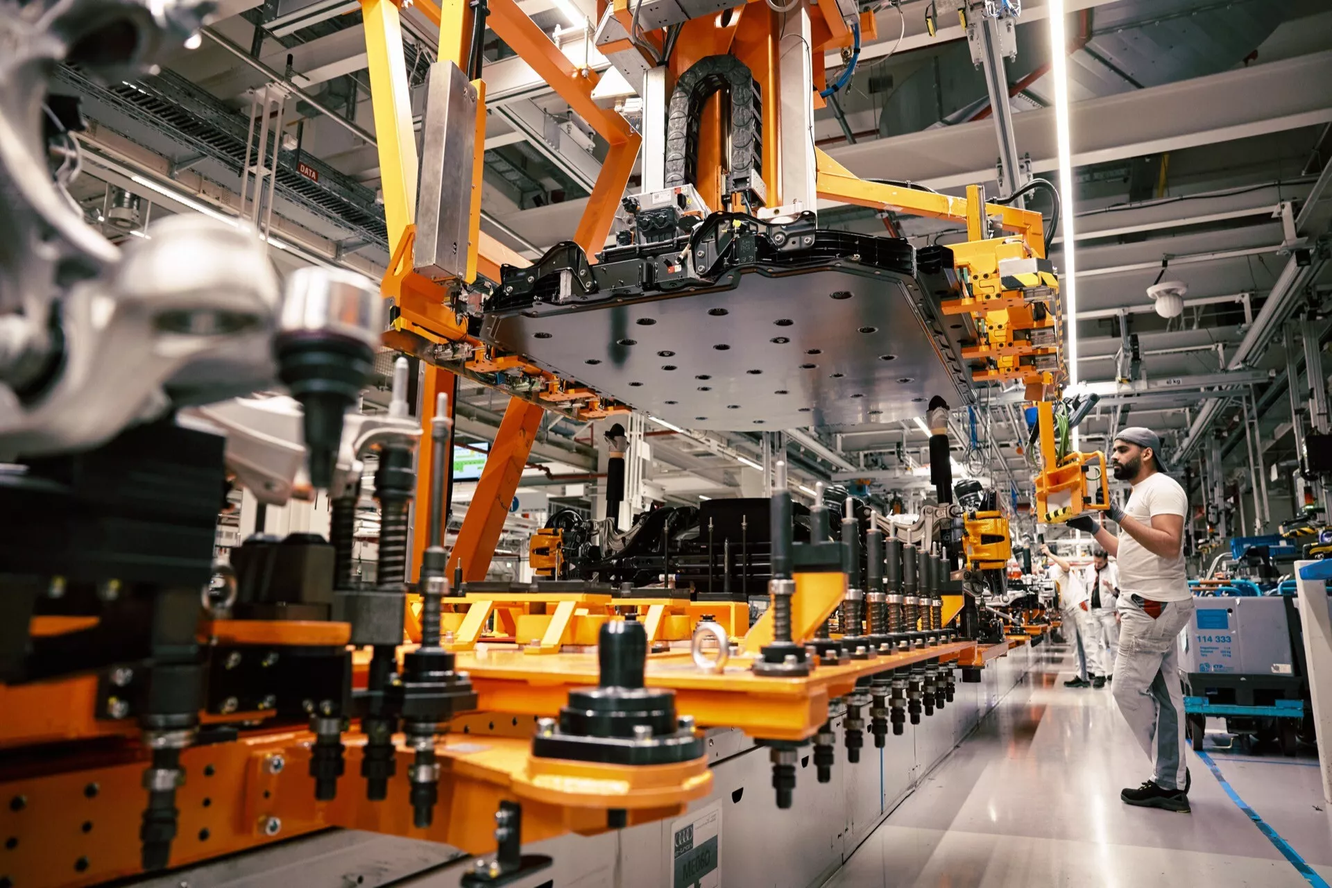 Montagem do Audi Q8 e-tron 2023 em uma planta industrial, mostrando um braço robótico manipulando o chassis monobloco do veículo sem portas ou rodas em uma linha de montagem automotiva