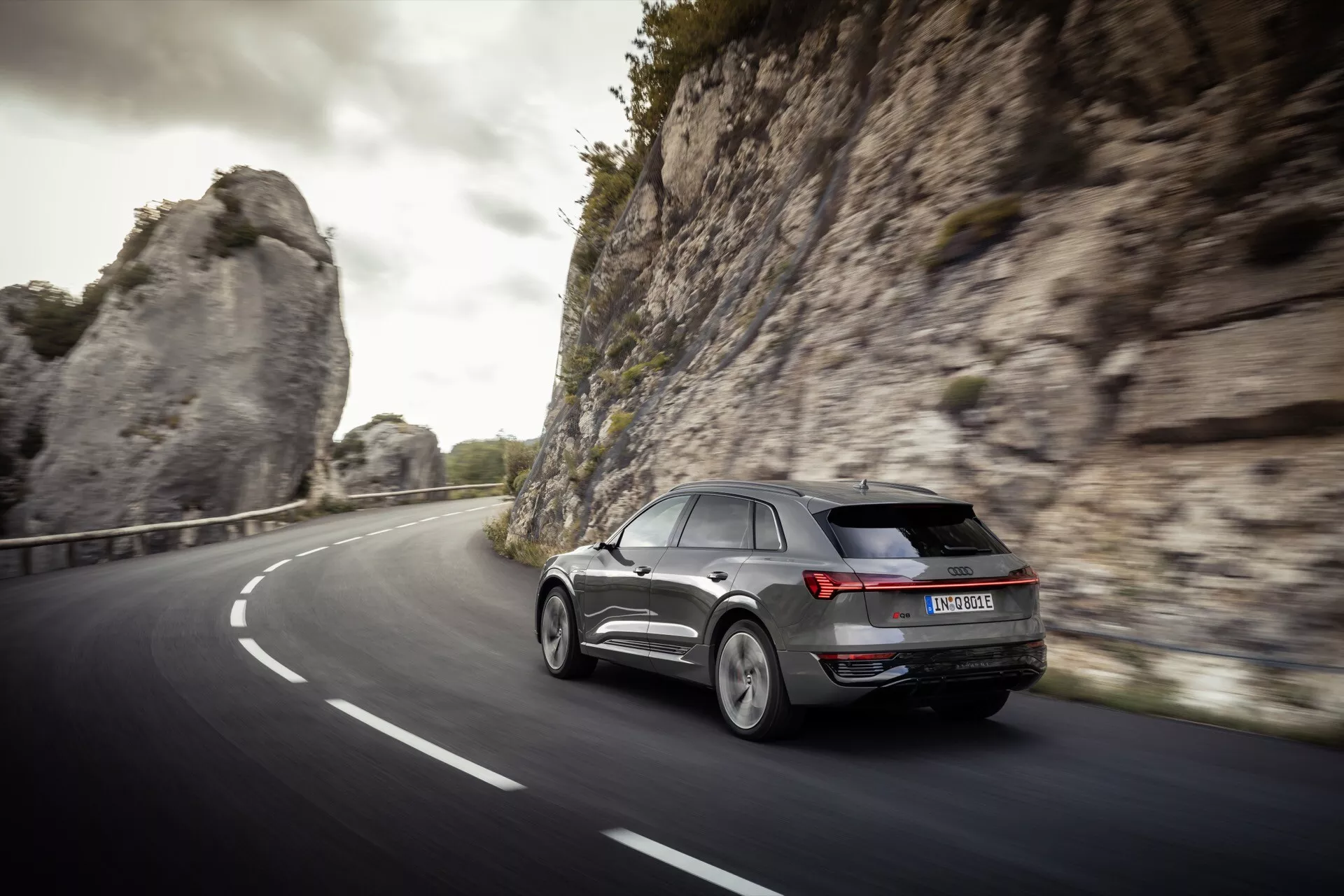 SUV moderno Audi Q8 e-tron 2023 em vista dinâmica traseira de três quartos dirigindo em uma estrada curva de duas pistas através de um ambiente natural, possivelmente montanhoso, mostrando luzes traseiras LED, rodas de liga leve e design aerodinâmico sob luz suave