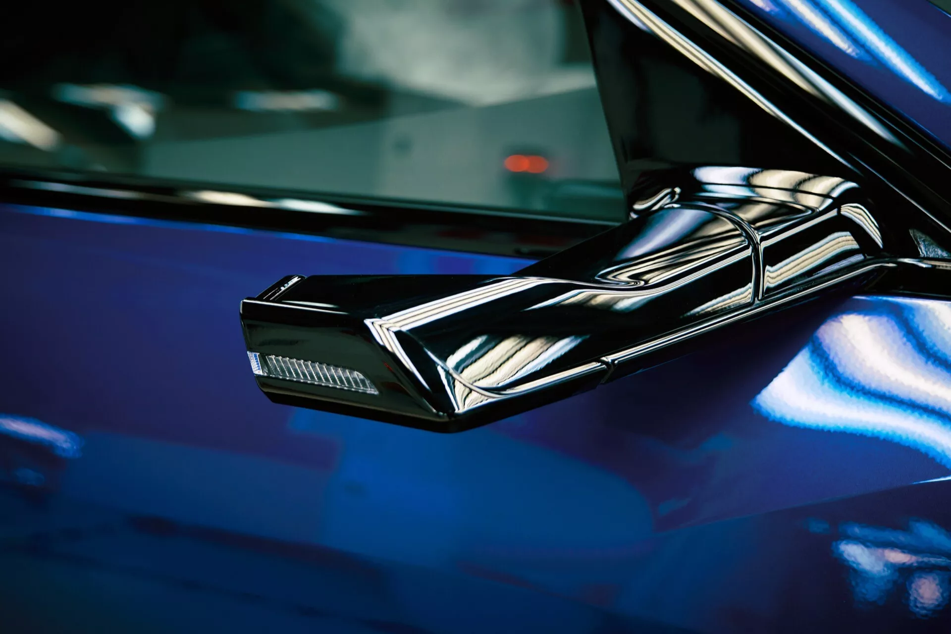 Close-up de um espelho lateral preto, elegante e angular de um Audi Q8 e-tron 2023, destacando elementos de design futurista e luzes de seta integradas contra uma superfície de carro azul profundo e brilhante
