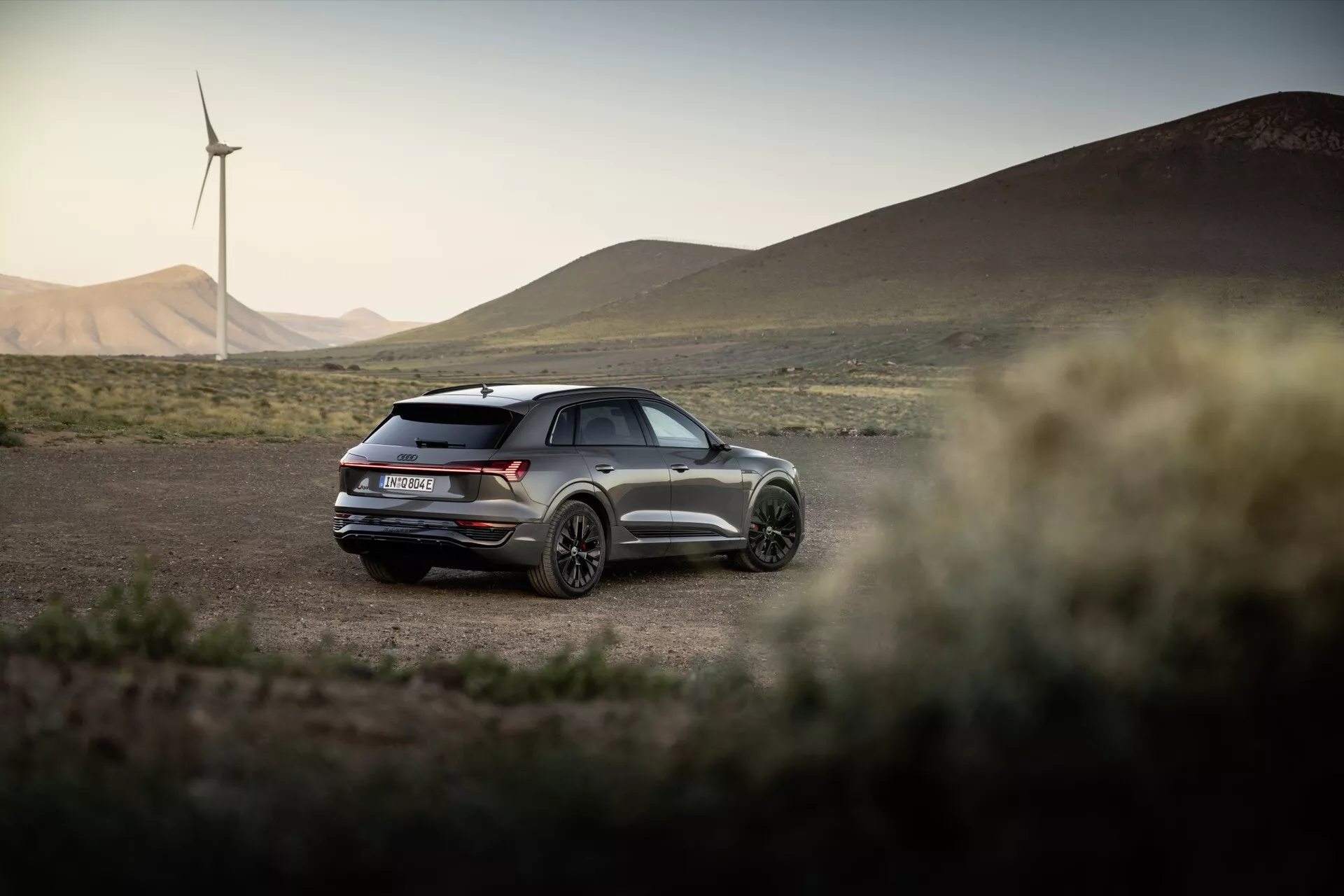 Audi Q8 e-tron 2023 em ambiente natural ao ar livre com turbinas eólicas ao fundo, exibindo seu design esportivo com teto panorâmico, escapamentos duplos e visão traseira de três quartos ao final da tarde