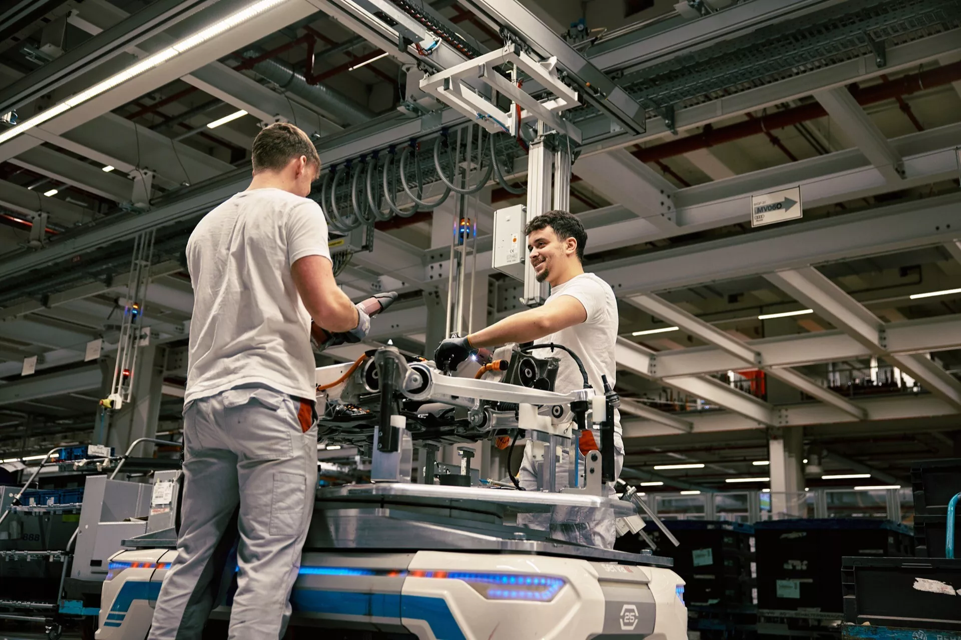 Ambiente industrial com dois trabalhadores montando ou mantendo equipamentos mecânicos no chão de fábrica, sob iluminação artificial, com um ponto de vista elevado enfatizando o ambiente de fabricação