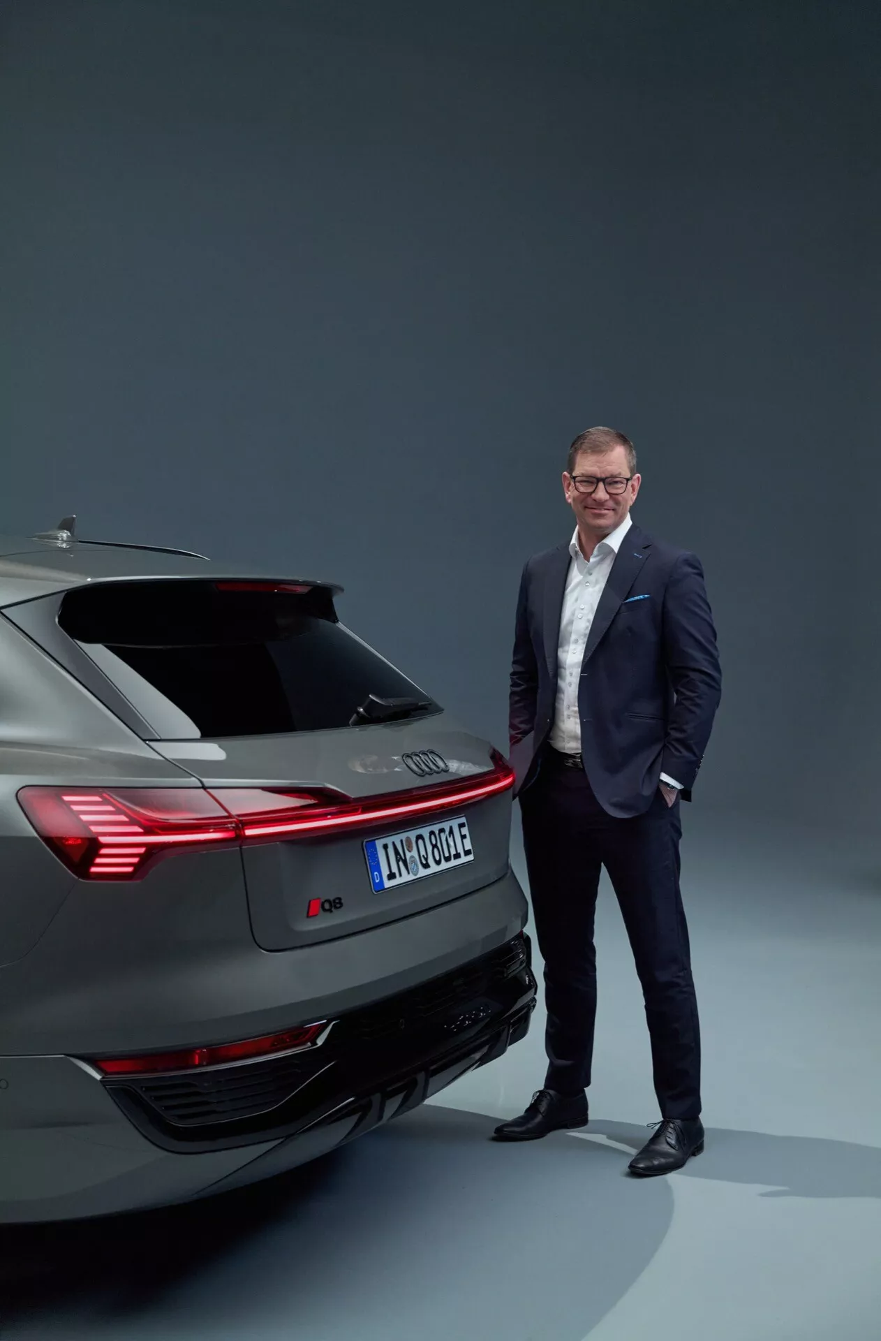 Veículo elétrico Audi Q8 e-tron 2023 mostrando design traseiro incluindo lanternas traseiras, para-choque e sistema de exaustão em um ambiente de estúdio, com um homem vestido profissionalmente ao lado, enfatizando tecnologia automotiva moderna e luxo.