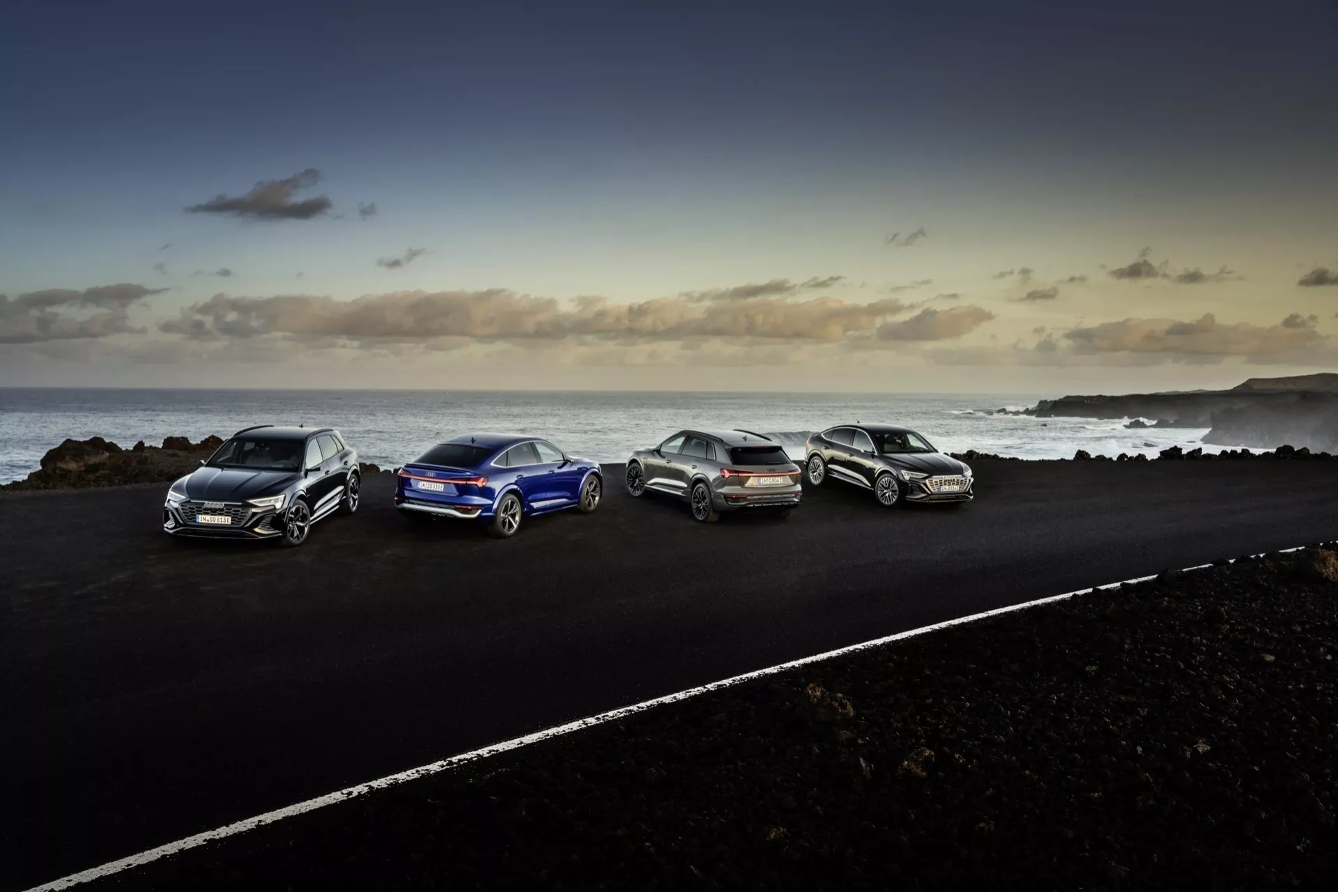 Audi Q8 e-tron 2023 estacionado em estrada asfaltada em ambiente costeiro com terreno rochoso e oceano ao fundo ao entardecer, exibindo design aerodinâmico, rodas de liga leve e iluminação moderna