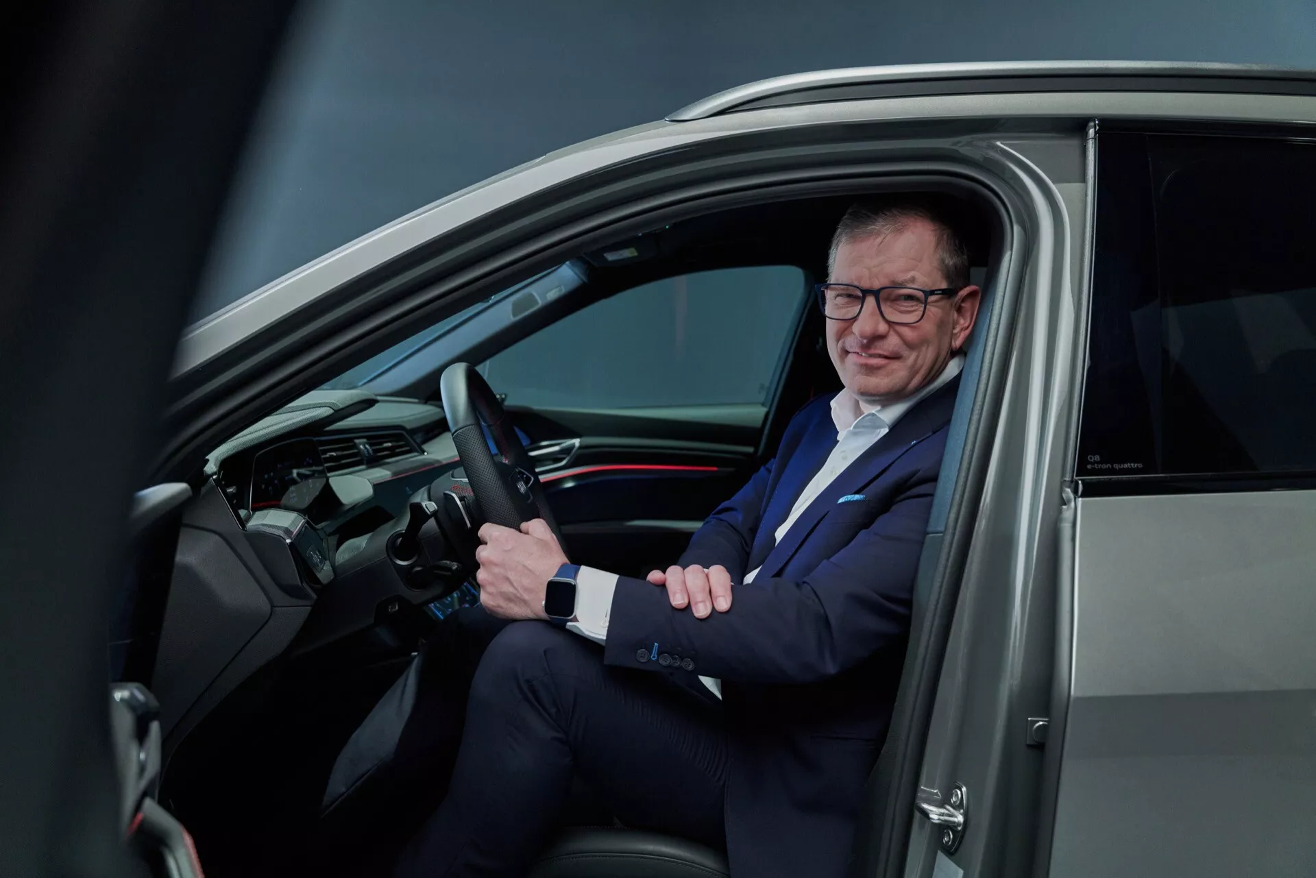 Vista interior do Audi Q8 e-tron 2023 através da porta do lado do motorista aberta mostrando o volante com controles montados, painel elegante com display digital, e um homem vestido profissionalmente no banco do motorista, refletindo a tecnologia moderna do veículo e um estilo de vida empresarial