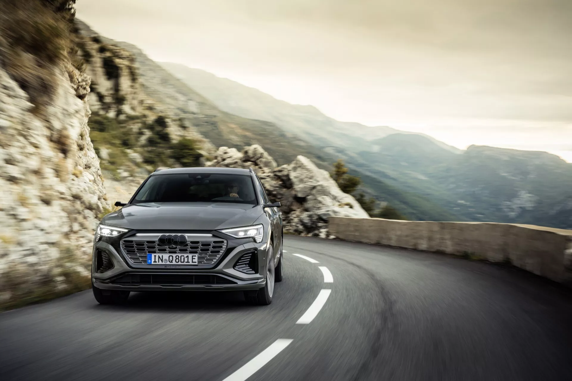 Audi Q8 e-tron 2023 em uma estrada sinuosa com montanhas rochosas ao fundo, apresentando design aerodinâmico e tecnologia avançada de faróis, transmitindo luxo e desempenho.