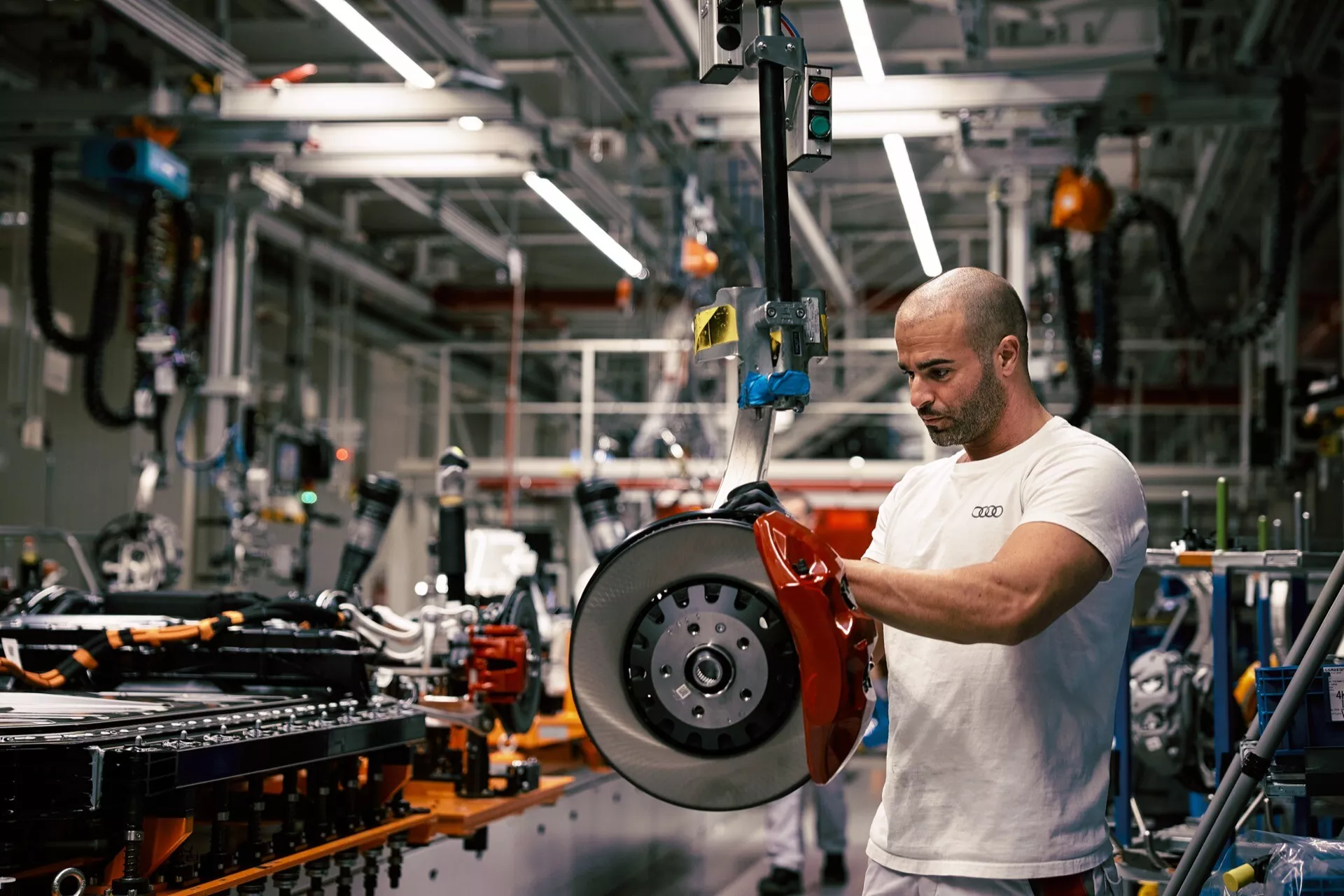 Vista de perto de um pinça de freio vermelho de alto desempenho e rotor de disco ventilado em um ambiente de produção automotiva, com um técnico trabalhando na montagem do componente em um chassis de veículo ao fundo