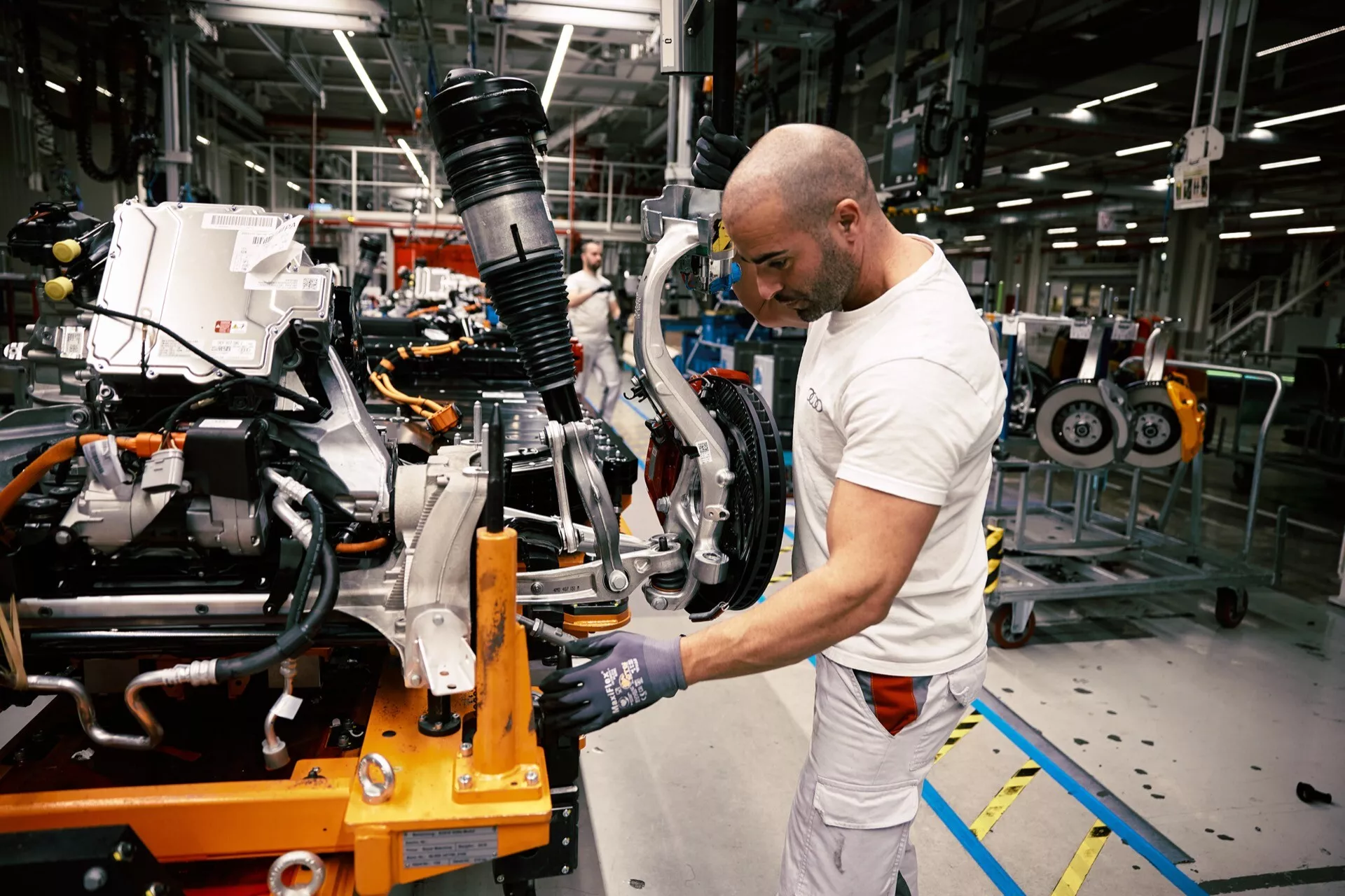 Chassi traseiro do Audi Q8 e-tron 2023 em estágio de montagem com suspensão e montagem de freio visíveis, e trabalhador utilizando ferramenta elétrica em fábrica de montagem de veículos
