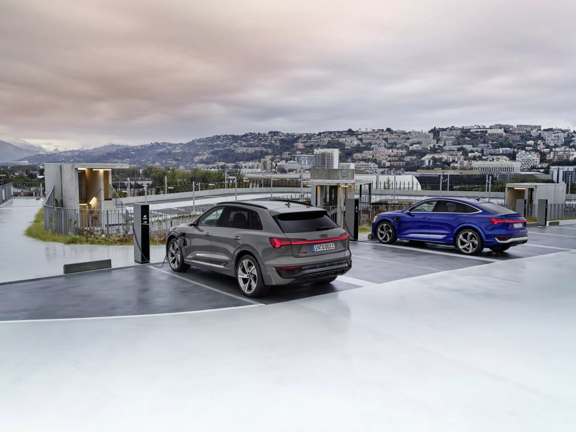 Audi Q8 e-tron 2023 em cinza e coupé esportivo em azul estacionados em um estacionamento de luxo no topo de um edifício ao amanhecer ou ao entardecer com paisagem urbana ao fundo, exibindo aerodinâmica avançada, design esportivo, lanternas traseiras LED, sistema de escapamento duplo e vidros escurecidos