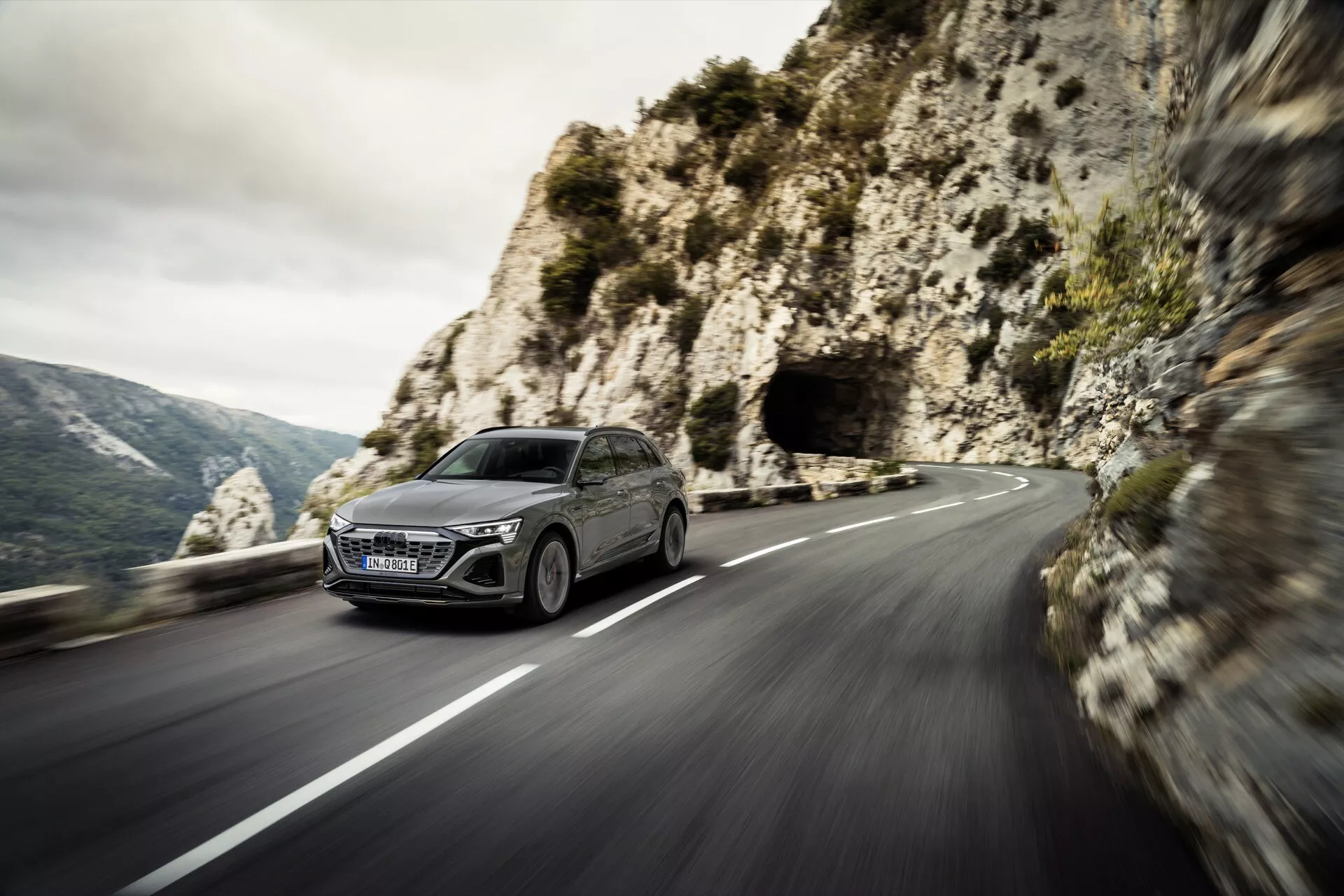 Audi Q8 e-tron 2023 navegando dinamicamente por uma estrada de montanha sinuosa, mostrando seu design moderno de hatchback, faróis LED e grade grande vista de um quarto frontal com um cenário montanhoso cênico