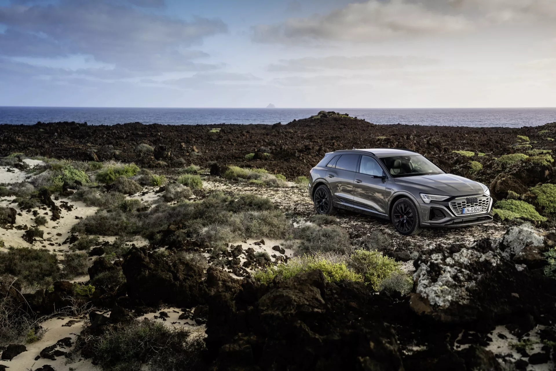 Audi Q8 e-tron 2023 em cinza metálico, exemplificando luxo com um design elegante e desportivo, capturado de um ângulo frontal lateral em terreno acidentado sob iluminação suave, destacando a tecnologia avançada de faróis LED, a grade proeminente com um grande emblema, contornos laterais dinâmicos e rodas de liga de múltiplos raios.