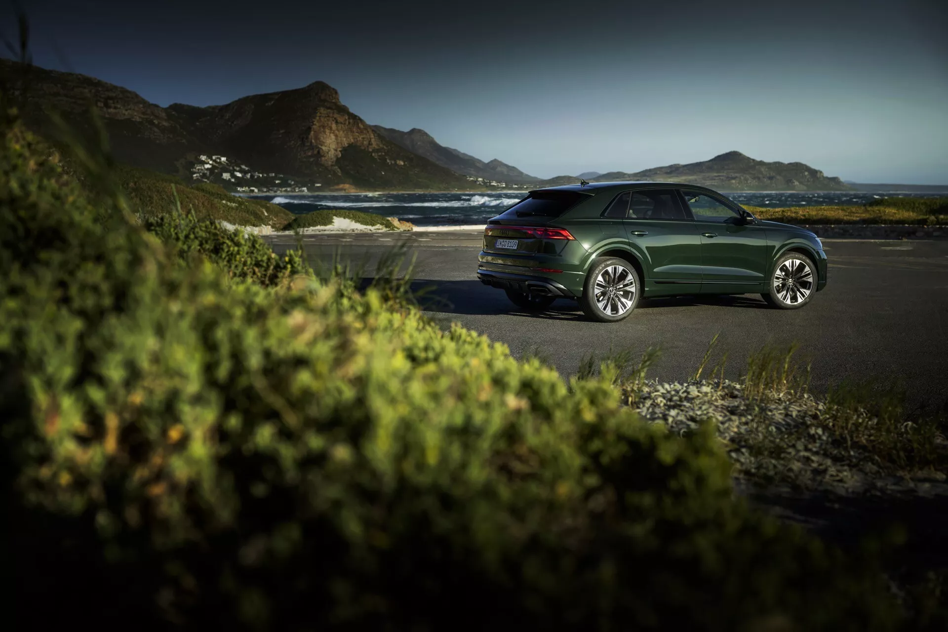 Sedan Audi Q8 (4M-Facelift) de cor verde-escuro estacionado em uma estrada costeira durante a hora dourada, exibindo linhas de design elegantes, rodas de liga leve multiraiadas e um ambiente externo sereno e luxuoso