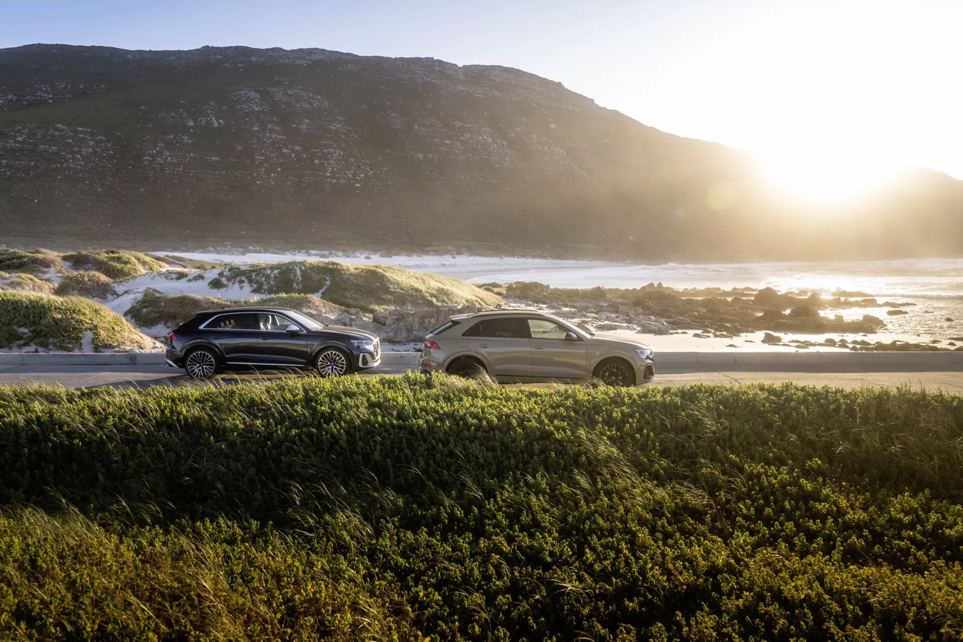 SUV e sedan Audi Q8 4M-Facelift 2023 estacionados durante o nascer ou pôr do sol com oceano e costa rochosa ao fundo, apresentando um brilho quente e reflexos de lente, destacando seus designs aerodinâmicos, luzes traseiras LED e superfícies reflexivas
