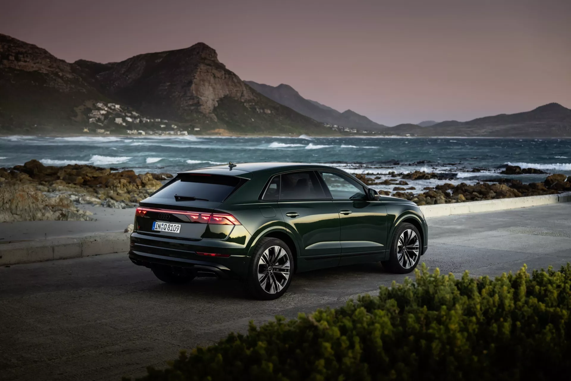 Audi Q8 2023 (4M-Facelift) estacionado em uma localização pitoresca à beira-mar ao amanhecer ou ao entardecer, exibindo sua vista traseira de três quartos, acabamento verde metálico escuro, e rodas de liga leve de múltiplos raios contra um tranquilo cenário montanhoso