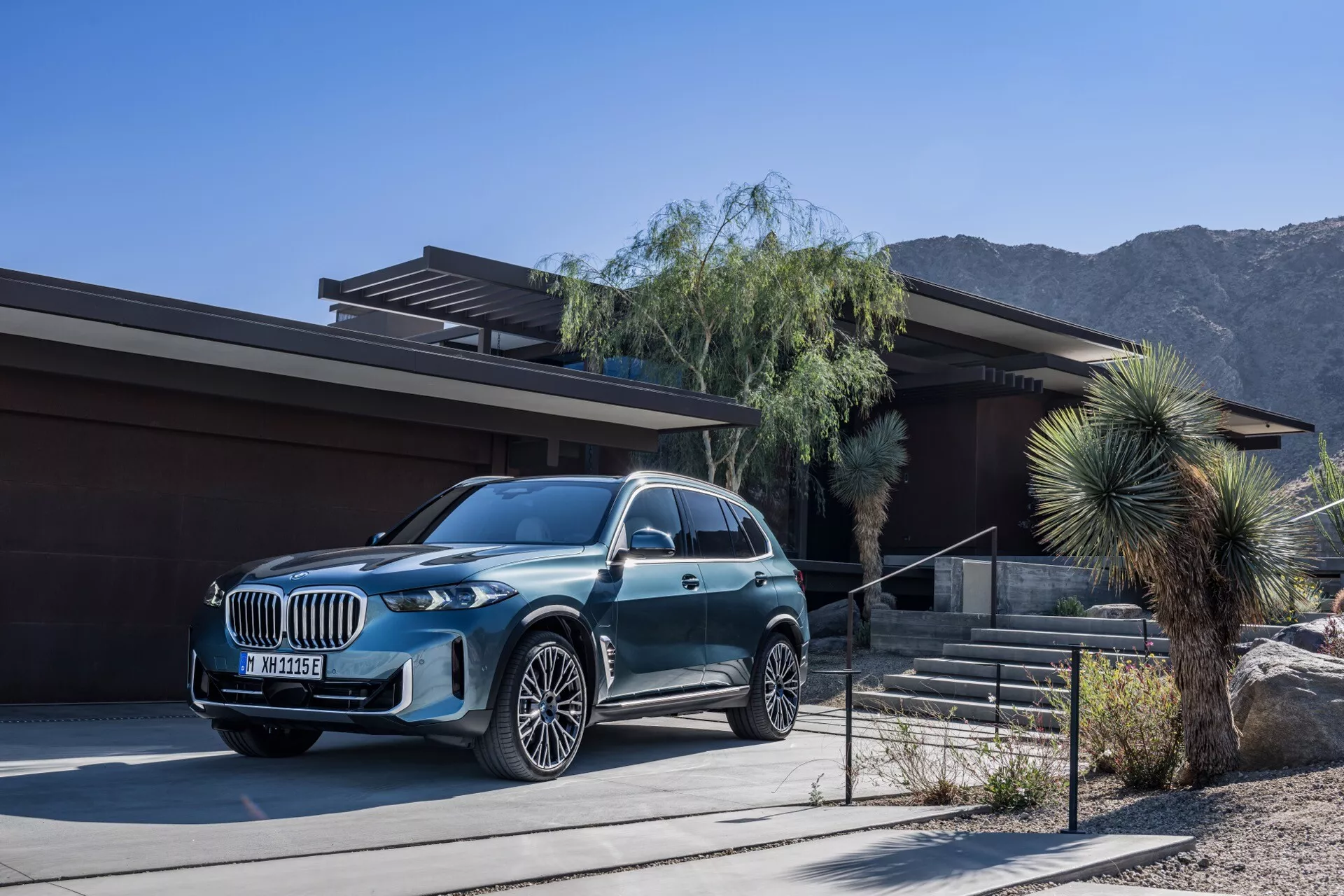SUV de luxo BMW X5 2023 (G05-F95 Facelift) estacionado em frente a uma casa contemporânea com uma paisagem de deserto e montanhas ao fundo, apresentando um acabamento metálico, grade grande, faróis LED e rodas de liga leve com vários raios