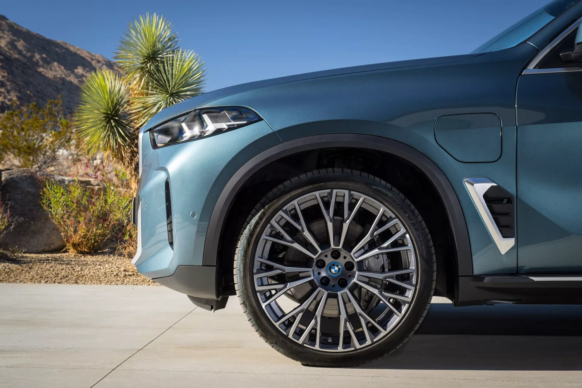 Imagem em close do BMW X5 (G05-F95) facelift 2023 destacando a roda dianteira e o paralama com suspensão avançada, roda de liga leve de padrão estrela em dois tons e pneu de baixo perfil orientado para desempenho, exibindo design aerodinâmico esportivo e tecnologia de assistência ao estacionamento com fundo desértico diurno.