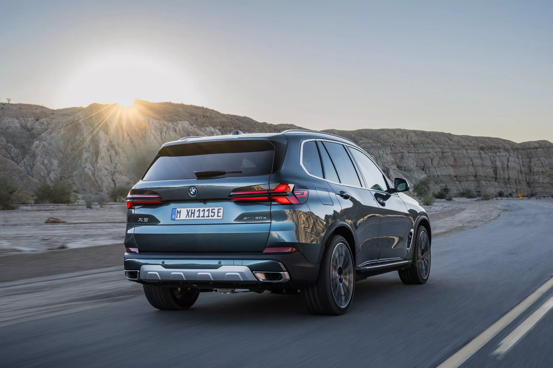 SUV moderno BMW X5 2023 (G05-F95-Facelift) em movimento em uma estrada cênica ao nascer ou pôr do sol, capturando ângulo traseiro dinâmico com luzes traseiras LED, barras de teto e características de tecnologia avançada sob um suave reflexo de luz solar.