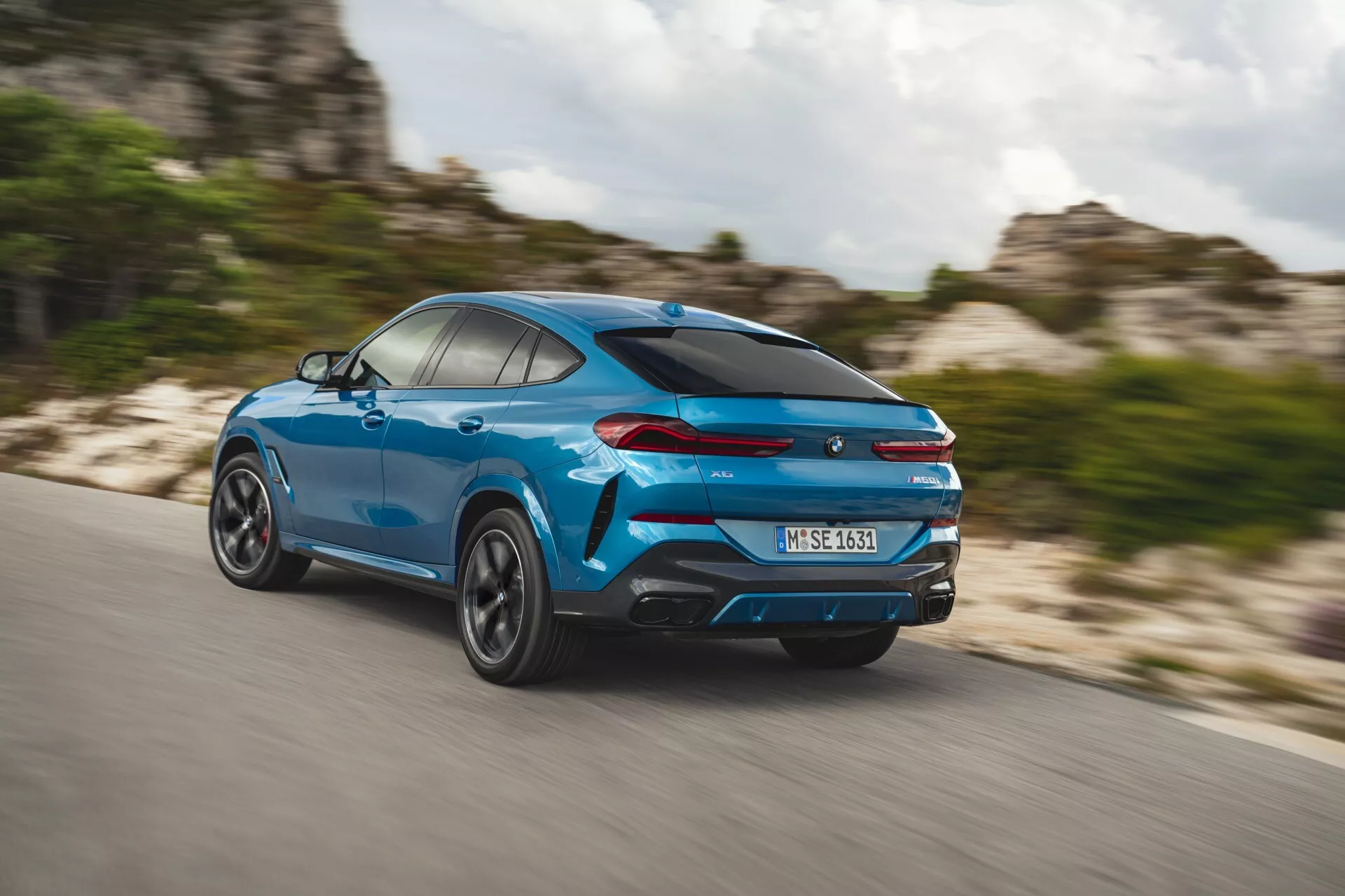 BMW X6 2023 (G06-F96-Facelift) em azul vibrante, capturado em vista traseira dinâmica de três quartos mostrando perfil lateral, rodas girando e design elegante de SUV estilo coupé com características de desempenho como escapamentos duplos e spoiler traseiro sob efeito de desfoque de movimento em uma rodovia.