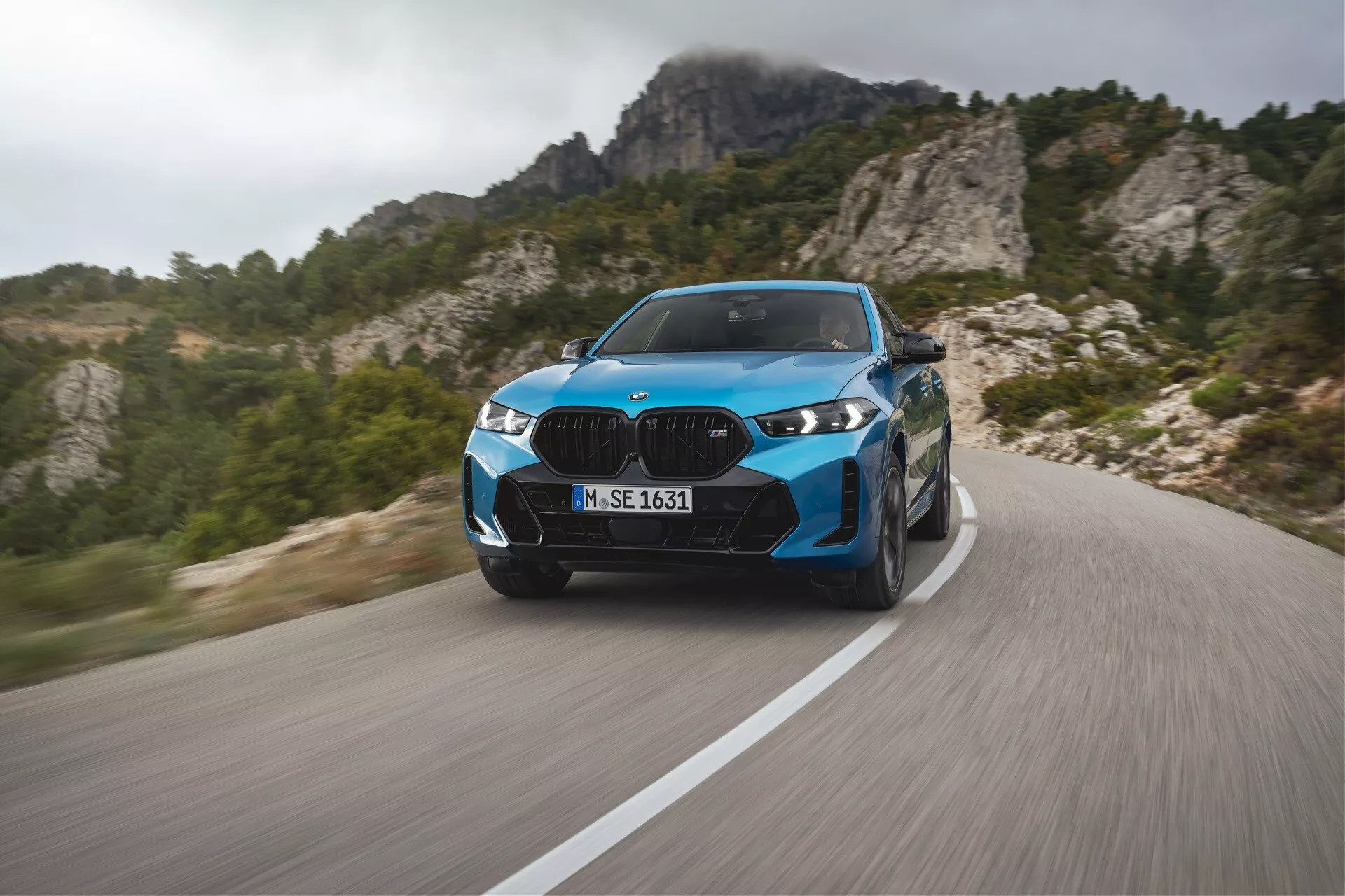 BMW X6 2023 (G06-F96-facelift) em azul vibrante, exibindo performance de luxo moderna em uma estrada montanhosa sinuosa com desfoque de movimento dinâmico, faróis LED avançados, elementos de design elegantes e rodas esportivas, contra um cenário de terreno montanhoso.