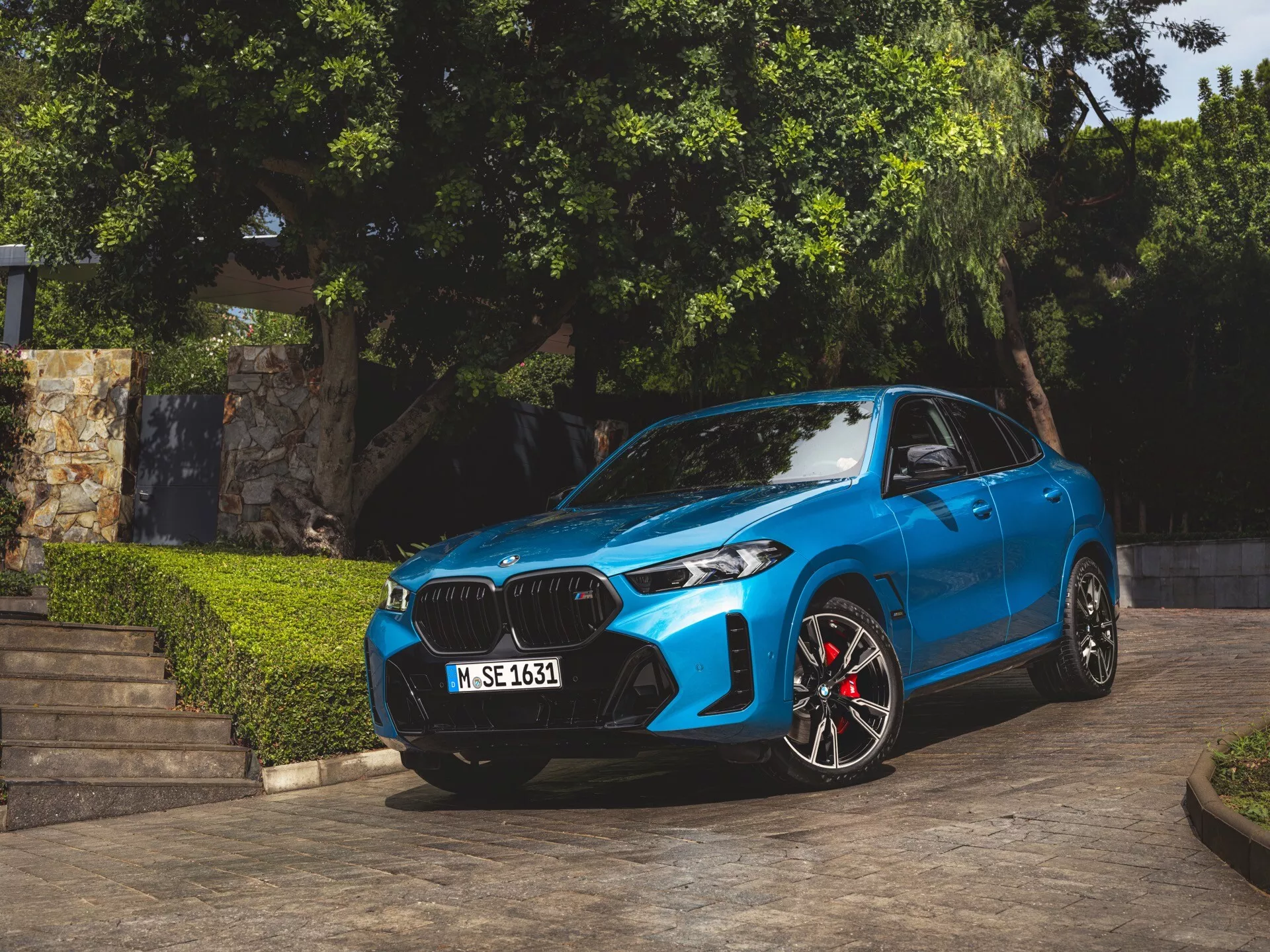 BMW X6 2023 (G06-F96-Facelift) em azul, estacionado em ângulo mostrando sua fachada frontal, grade em forma de rim, faróis LED ou laser, rodas esportivas de liga leve com pinças de freio vermelhas, linhas contornadas no capô e espelhos laterais aerodinâmicos com setas integradas, capturado à luz natural do dia com vegetação e degraus arquitetônicos ao fundo