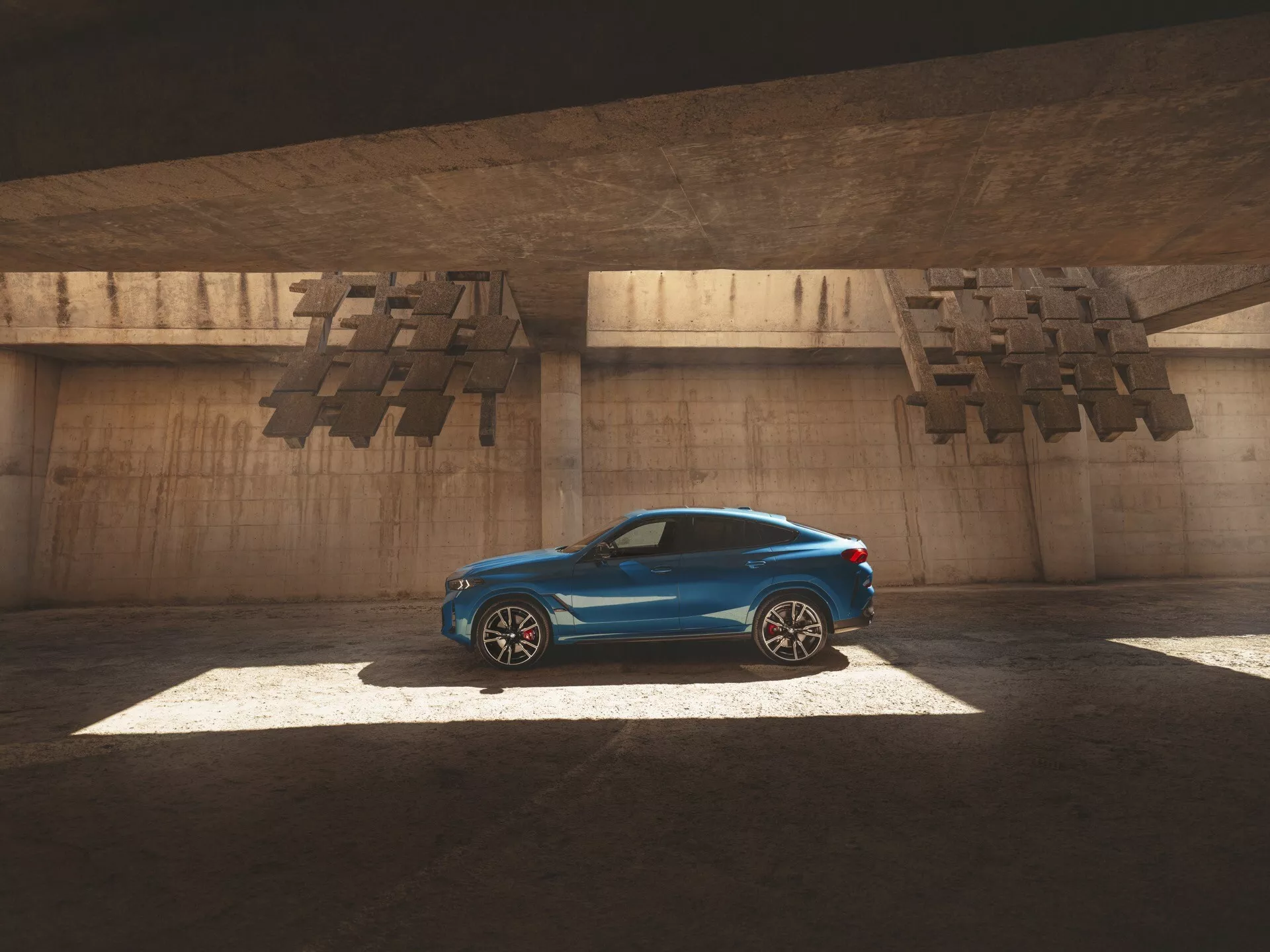 Carro estilo coupé BMW X6 2023 (G06-F96 Facelift) em azul brilhante, destacado pela luz do sol através de uma abertura acima, capturando seu perfil lateral elegante, linhas aerodinâmicas e características de luxo como rodas de liga leve e teto solar