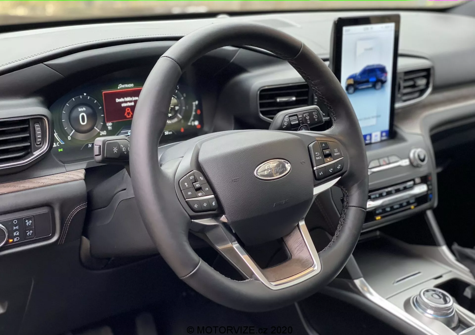 Vista do interior do Ford Explorer 2019 a partir da perspectiva do assento do motorista, com um moderno volante de três raios com acento cromado e controles multifuncionais para controle de cruzeiro, mídia e comandos de voz. Atrás do volante encontra-se um painel de instrumentos digital exibindo informações do veículo. O painel inclui uma grande tela sensível ao toque inclinada em direção ao motorista para navegação e acesso a mídia, complementada por um seletor de marchas rotativo e materiais de alta qualidade.
