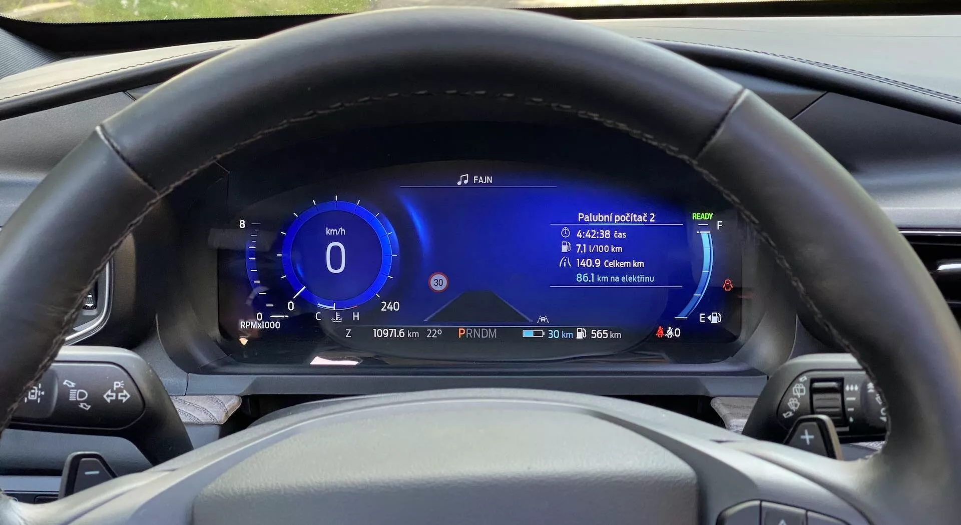 Visão interna de um Ford Explorer 2019, a partir da perspectiva do assento do motorista, focando no volante central e no painel digital atrás dele. O volante possui vários botões de controle em ambos os raios. Atrás dele, um painel de instrumentos totalmente digital exibe informações como velocidade, medidor de potência, alcance de condução, nível de combustível, temperatura externa e leitura de eficiência. O painel também exibe a hora e a data, e é projetado com elementos modernos e tecnologicamente avançados em azul e branco. O volante é feito de couro preto com detalhes em costura.