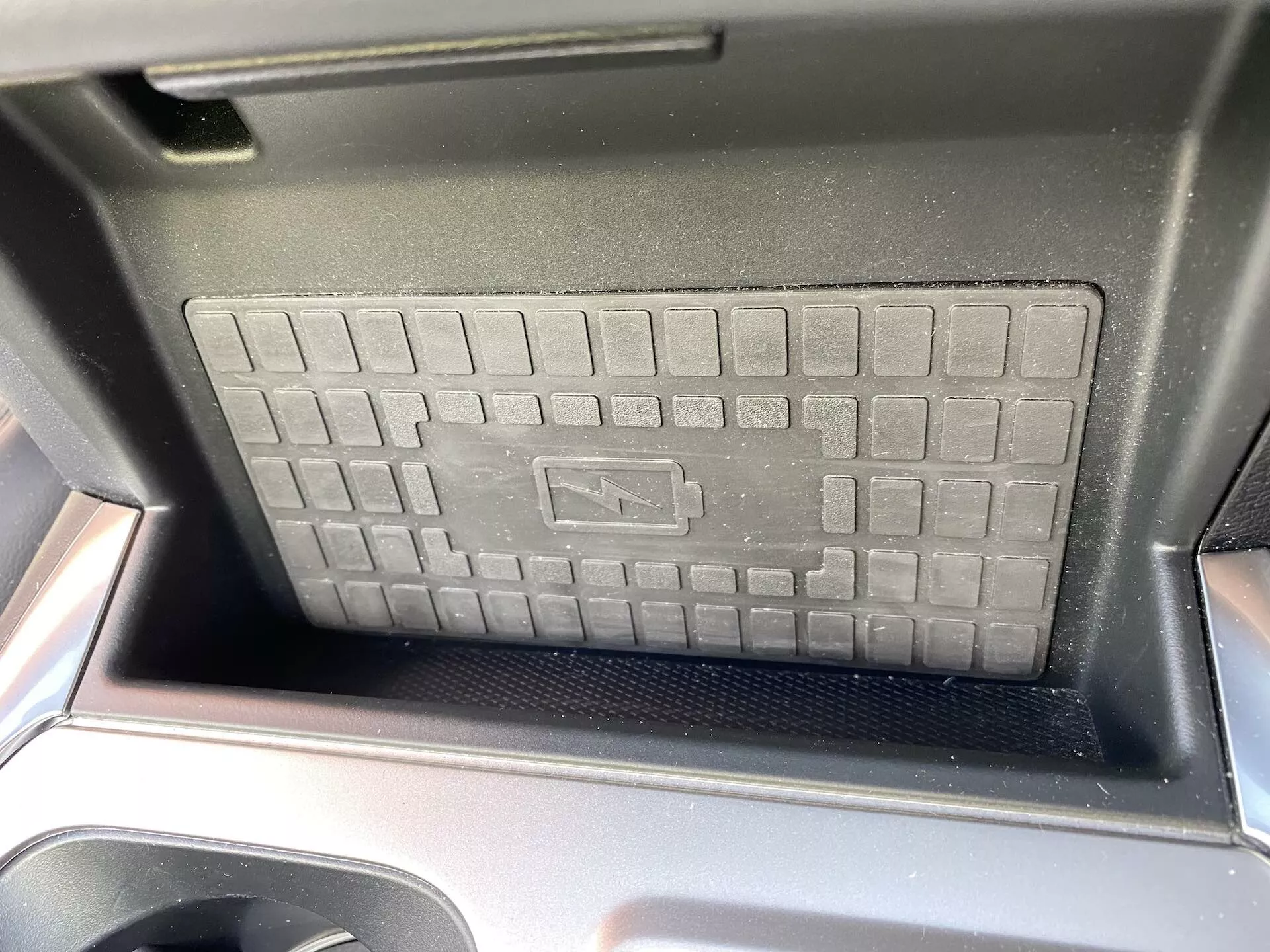 Close-up do compartimento do console central do Ford Explorer 2019, destacando o carregador sem fio com textura elevada em forma de grade e superfície antiderrapante. Bordas em plástico preto brilhante são visíveis, adicionando um toque de design automotivo moderno. A luz solar sugere uma foto tirada durante o dia a partir do assento dianteiro, com o símbolo de carregamento indicando tecnologia avançada de carregamento sem fio.