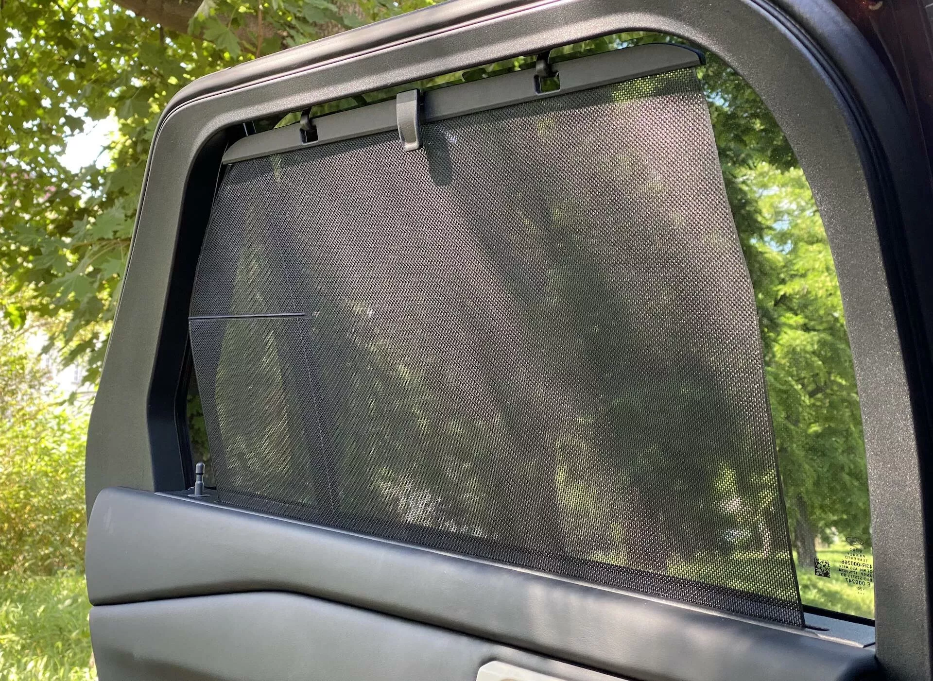 Vista interna aproximada da porta lateral traseira e vidro do Ford Explorer 2019. A imagem captura um vidro parcialmente abaixado com uma tela de proteção solar de malha instalada, cobrindo toda a área de vidro e se estendendo ligeiramente além da borda superior. A parte superior do painel da porta do carro, com um acabamento liso, também está visível. Tirada na orientação horizontal, a foto mostra a luz filtrando através da tela de proteção solar, sugerindo um ambiente externo parecido com um parque. Foco no conforto dos passageiros com acessório de proteção solar.