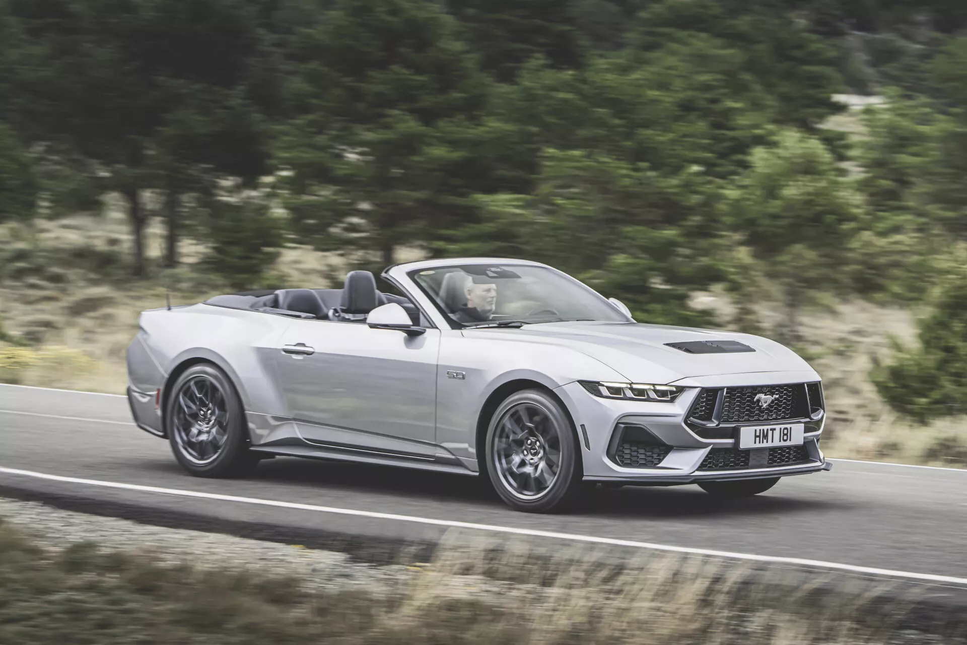 Ford Mustang Conversível 2024 em prata, exibindo seu estilo agressivo e recursos avançados em movimento numa rodovia, capturado de um ângulo de três quartos do lado do motorista