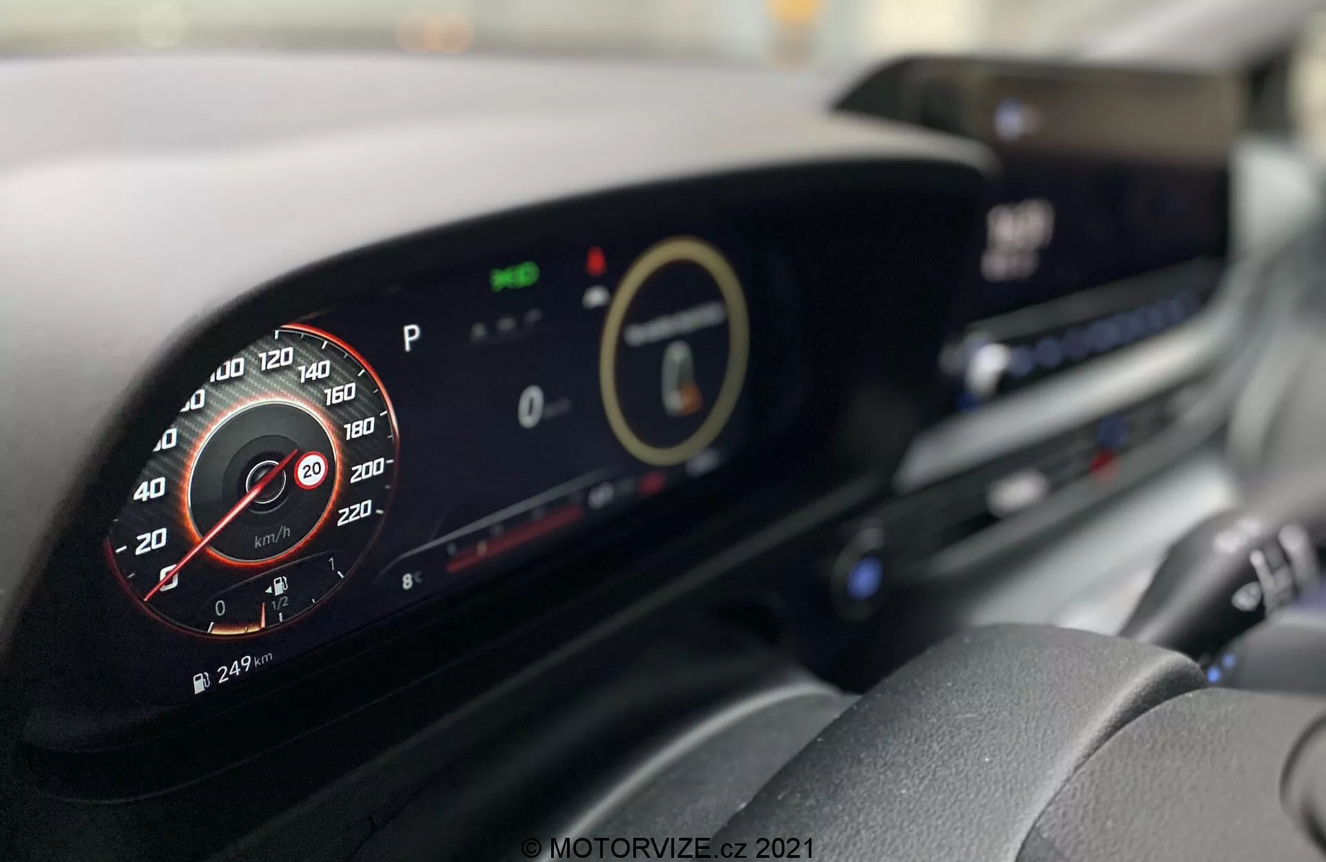 Interior do Hyundai i20 2020 com um painel moderno. Imagem tirada do assento do motorista com foco no painel de instrumentos digital e na tela central, mostrando detalhes como o tacômetro analógico simulado, indicadores ecológicos e um sistema de infoentretenimento. Características adicionais como saídas de ar e botões de controle do clima também são visíveis.
