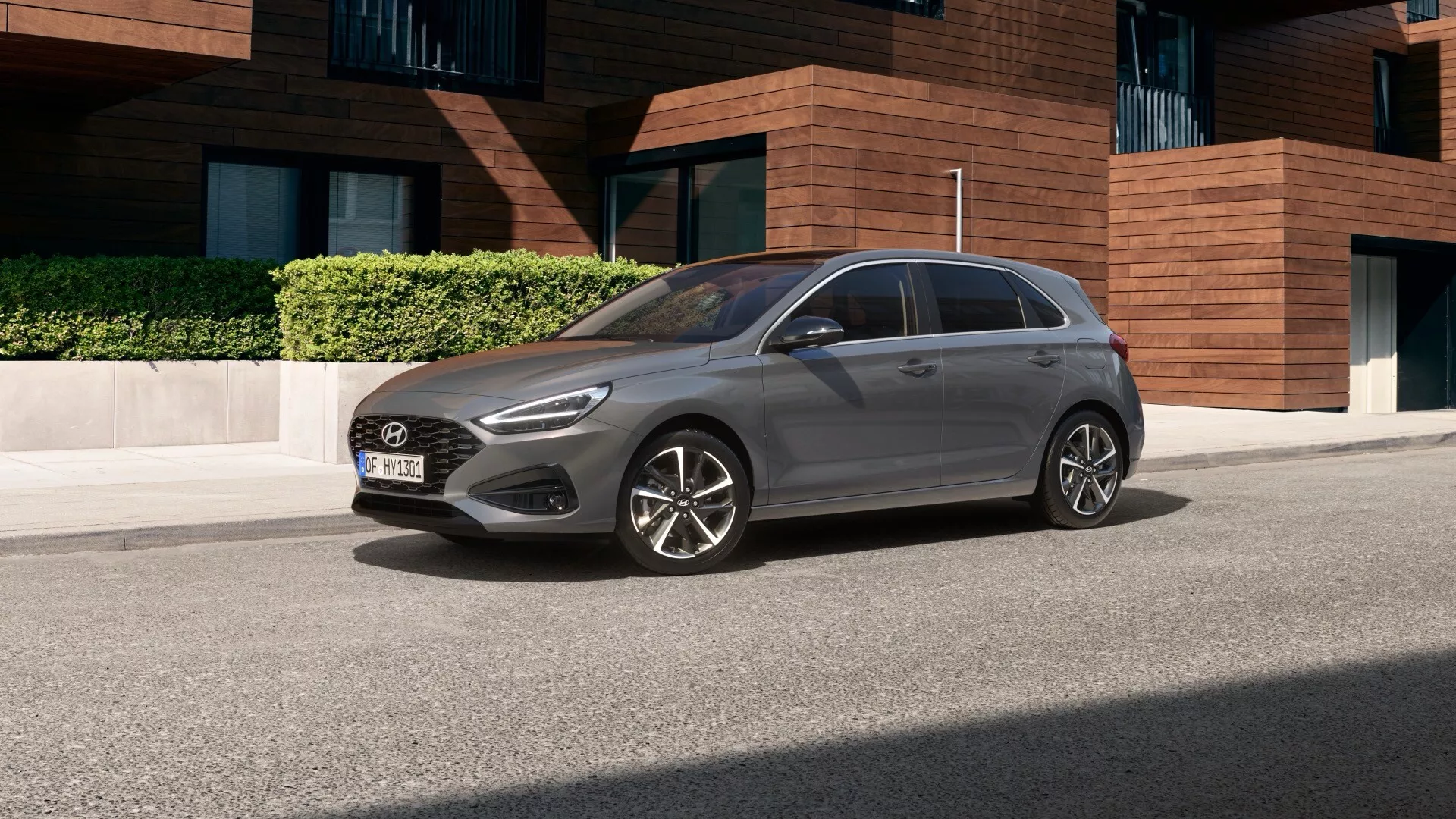 Um Hyundai i30 2024 fotografado de um ângulo ligeiramente elevado, capturando seu design geral, teto e parte do capô. O carro está estacionado em uma área residencial com arquitetura moderna ao fundo, mostrando um hatchback de quatro portas com rodas de liga leve, sugerindo um nível de acabamento médio ou superior. O perfil lateral do lado do motorista é totalmente visível, destacando sua forma aerodinâmica, janelas escurecidas e antena no teto, com o entorno proporcionando uma visão clara de seu design estiloso e contemporâneo.