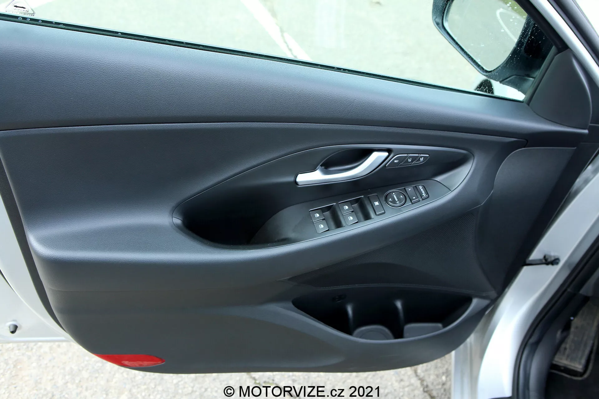 Painel da porta interior do Hyundai i30 2020 (i30-kombi) visto de um ângulo oblíquo, mostrando a maçaneta da porta com acabamento cromado prateado, botões de controle ergonômicos para os vidros elétricos, botão eletrônico de trava da porta, bolso de armazenamento e acabamento da porta com dupla textura, com painel superior macio e parte inferior de plástico rígido em cores escuras.