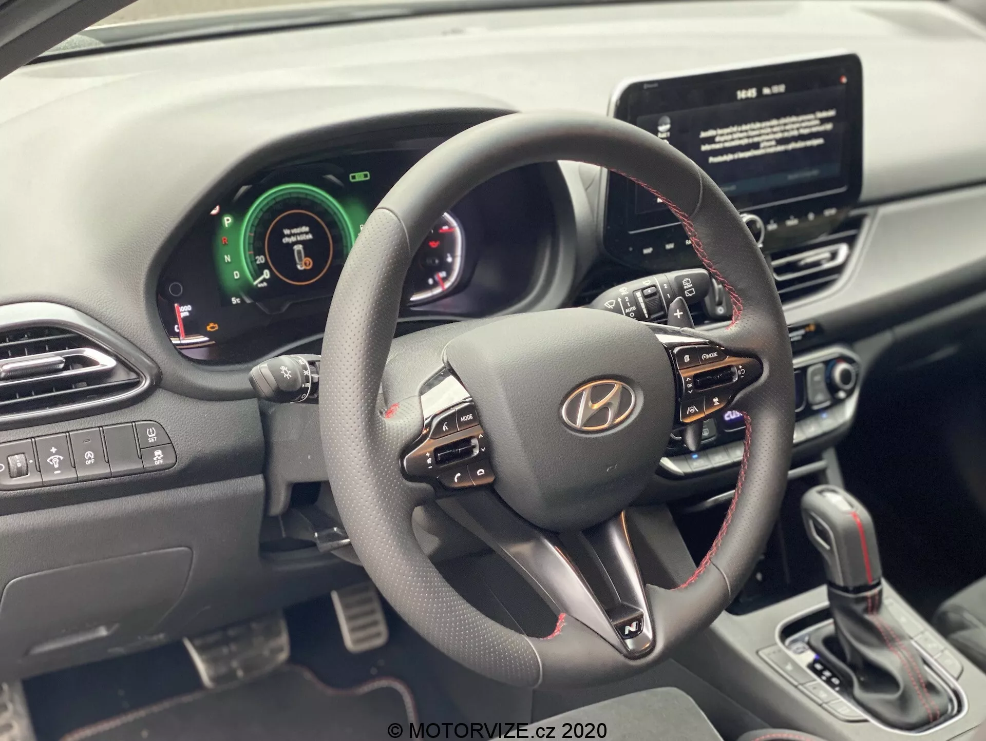Vista interior de um Hyundai i30 2020 (i30-kombi) a partir do banco do motorista, focando no volante e painel de instrumentos. O volante moderno possui múltiplos botões de controle para volume, comandos de voz e chamadas em viva-voz. O cluster de medidores combina elementos digitais e analógicos, indicando sistemas avançados de assistência ao motorista. Uma tela sensível ao toque central de infotainment é visível, sugerindo navegação e reprodução de mídia. O console central mostra botões de controle de clima e uma alavanca de câmbio para transmissão automática. Os controles de vidros elétricos e espelhos laterais estão na porta do motorista.