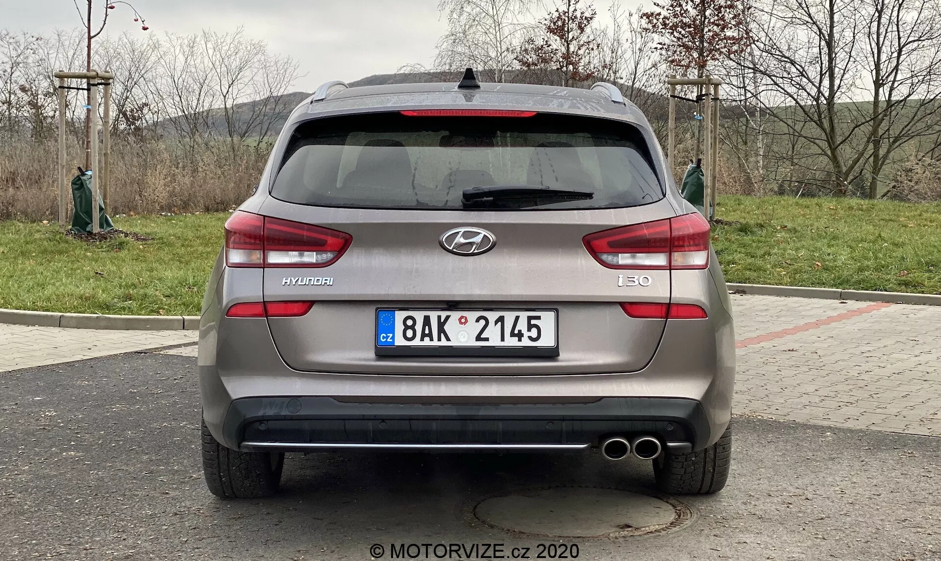 A imagem mostra a vista traseira de um Hyundai i30 Kombi 2020. O carro está estacionado e a foto é tirada de um ângulo direto, aproximadamente na altura do porta-malas do veículo. Centralizado no veículo, a imagem fornece uma visão clara e simétrica das características traseiras do carro, incluindo o logotipo da Hyundai, o nome do modelo 'i30' e o nível de acabamento 'CRDi', indicando um motor diesel de injeção direta Common Rail. Os detalhes visíveis incluem a placa de estilo europeu, luzes traseiras horizontais, sensores traseiros, ponteiras duplas de escape, limpador de para-brisa traseiro, antena em forma de barbatana de tubarão e possivelmente uma câmera traseira próxima à área da placa.