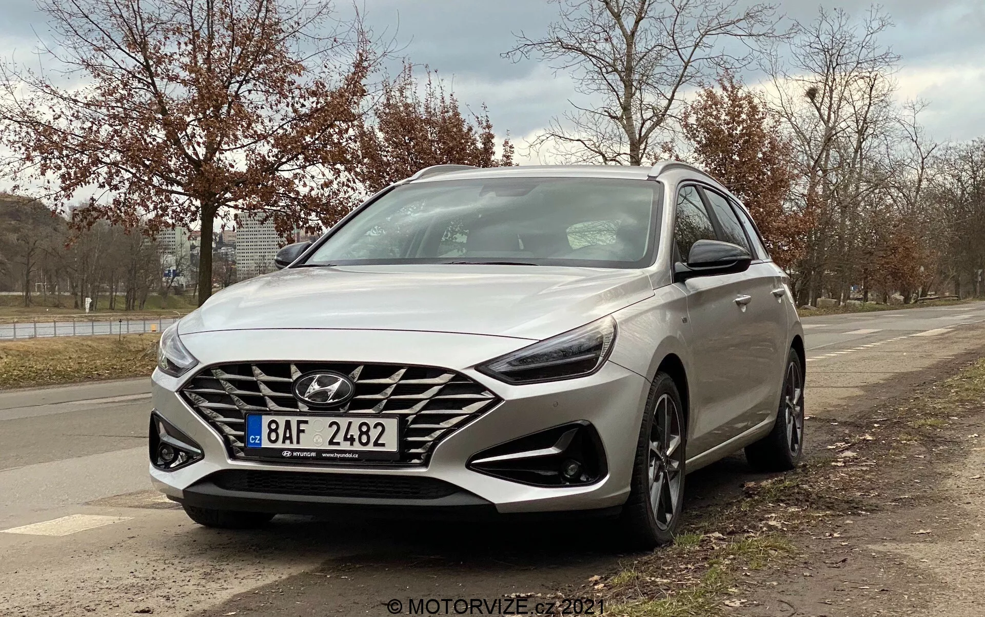 A imagem apresenta um Hyundai i30 Kombi 2020 prateado capturado de um ângulo baixo de três quartos da frente, mostrando o design do lado do motorista. O ângulo enfatiza a grande grade em cascata com um proeminente logotipo da Hyundai, faróis LED modernos, rodas de liga leve de vários raios, luzes diurnas integradas abaixo dos faróis e um sensor de radar visível para funções de segurança. As linhas e contornos aerodinâmicos do carro também são destacados, com espelhos laterais que integram sinais de seta. O fundo inclui um meio-fio, árvores e um céu nublado, sugerindo um ambiente externo suburbano.