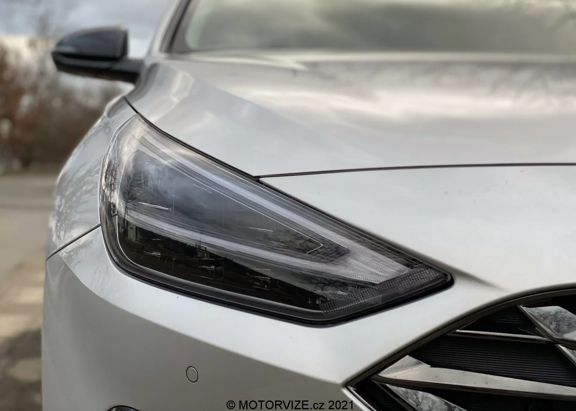 Imagem aproximada do Hyundai i30 2020 Kombi, mostrando a seção frontal esquerda do veículo em um ângulo de três quartos voltado para o lado do motorista, com foco no conjunto de faróis elegantes e angulares com tecnologia moderna LED ou HID, acompanhado por um espelho lateral aerodinâmico com possíveis integrações tecnológicas avançadas, como ajuste elétrico e monitoramento de ponto cego. A carroceria do carro exibe um acabamento perolado ou metálico de alta qualidade com contornos aerodinâmicos no capô e um indício da grade frontal. O fundo está desfocado para enfatizar os detalhes do carro.