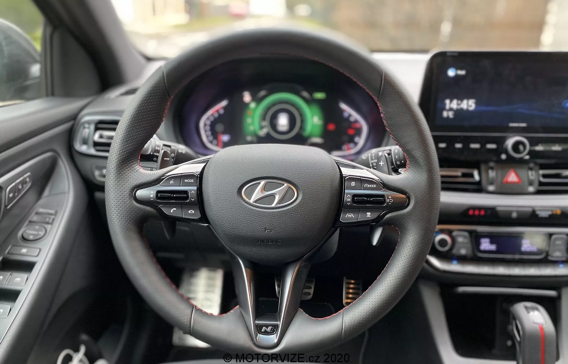 Vista da perspectiva do motorista do volante e do painel do Hyundai i30 2020, mostrando um design de três raios revestidos em couro com controles multifuncionais, um logotipo Hyundai proeminente e um cluster de instrumentos com mostradores analógicos e digitais. A tela sensível ao toque central do sistema de infoentretenimento e os botões de controle do clima destacam a tecnologia avançada do carro e os elementos de interação do motorista.