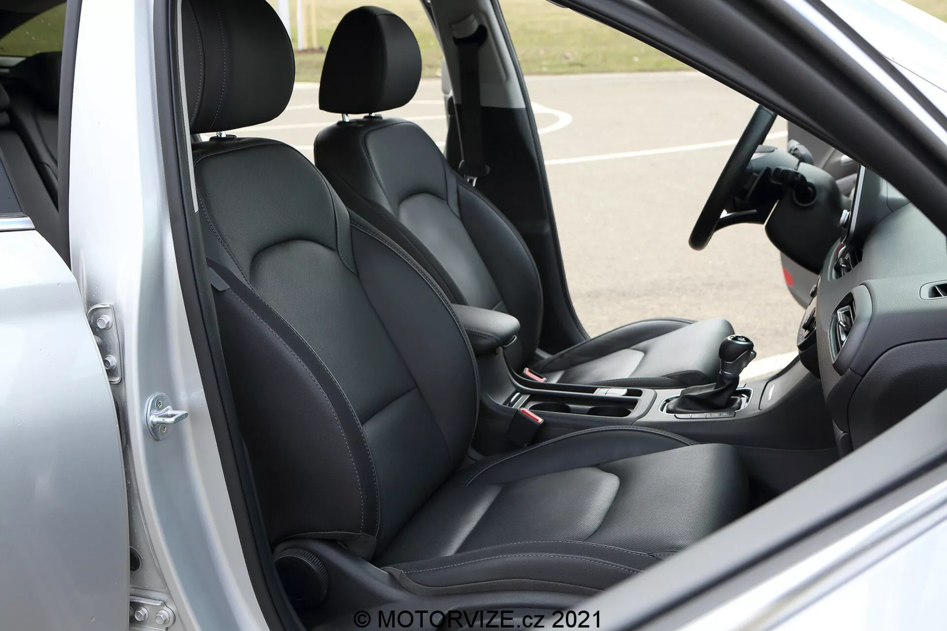 Vista lateral de close-up da cabine dianteira do Hyundai i30 Kombi 2020. A imagem mostra o assento do motorista e parte do banco do passageiro, ambos estofados em preto com um design contornado para maior conforto e suporte. Visível do lado do motorista com a porta aberta, a cena inclui uma alavanca de câmbio, freio de mão manual e parte dos sistemas de infotainment e controle climático, sugerindo recursos tecnológicos modernos. O painel da porta do motorista, mostrando os controles de vidros elétricos e possivelmente os ajustes do espelho, adiciona ao design funcional geral. O volante, embora parcialmente cortado, sugere controles integrados para melhor interação veicular. Marca d'água com 'MOTORVIZE.CZ 2021' no canto inferior direito.