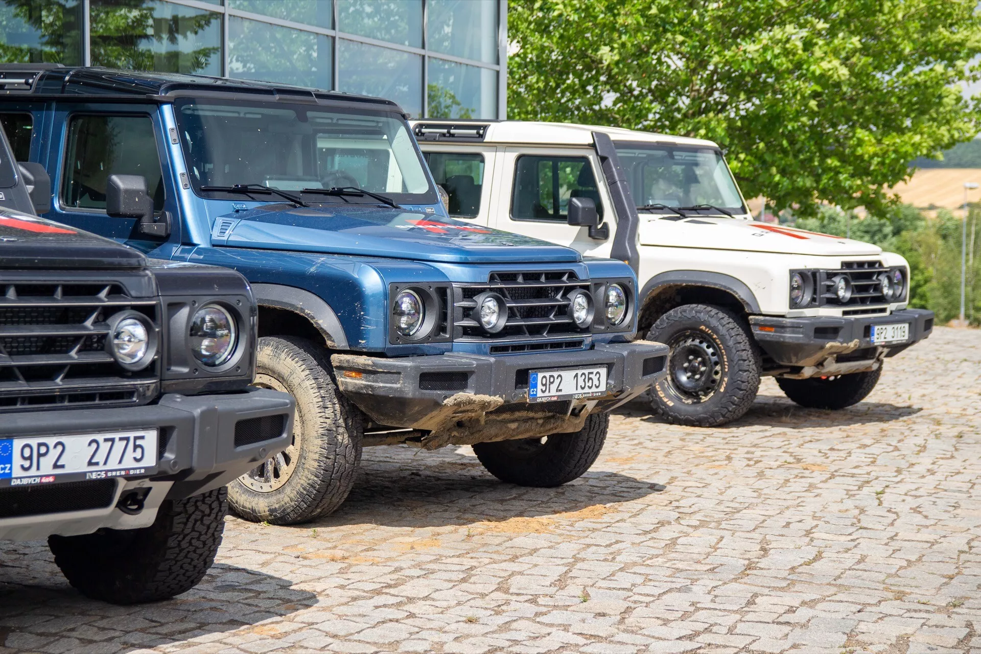 Formação da primeira geração do Ineos Grenadier 2022, com três SUVs com acessórios off-road estacionados num estacionamento, exibindo a grade frontal, faróis e design robusto e quadrado
