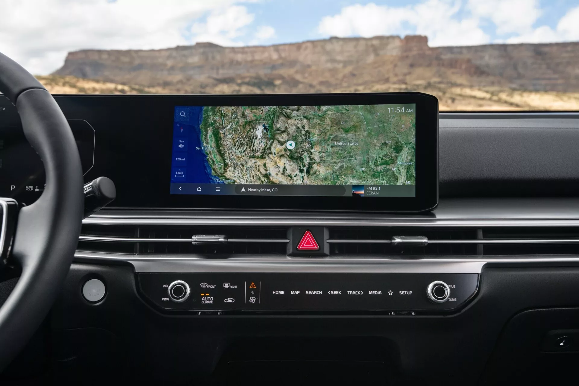 Vista do painel interior do Kia Sorento MQ4 facelift 2024 com display de infotainment touchscreen amplo com navegação GPS, controles manuais e sensíveis ao toque para clima e áudio, botão de luz de emergência e design minimalista com saídas de ar integradas, vista da perspectiva do motorista