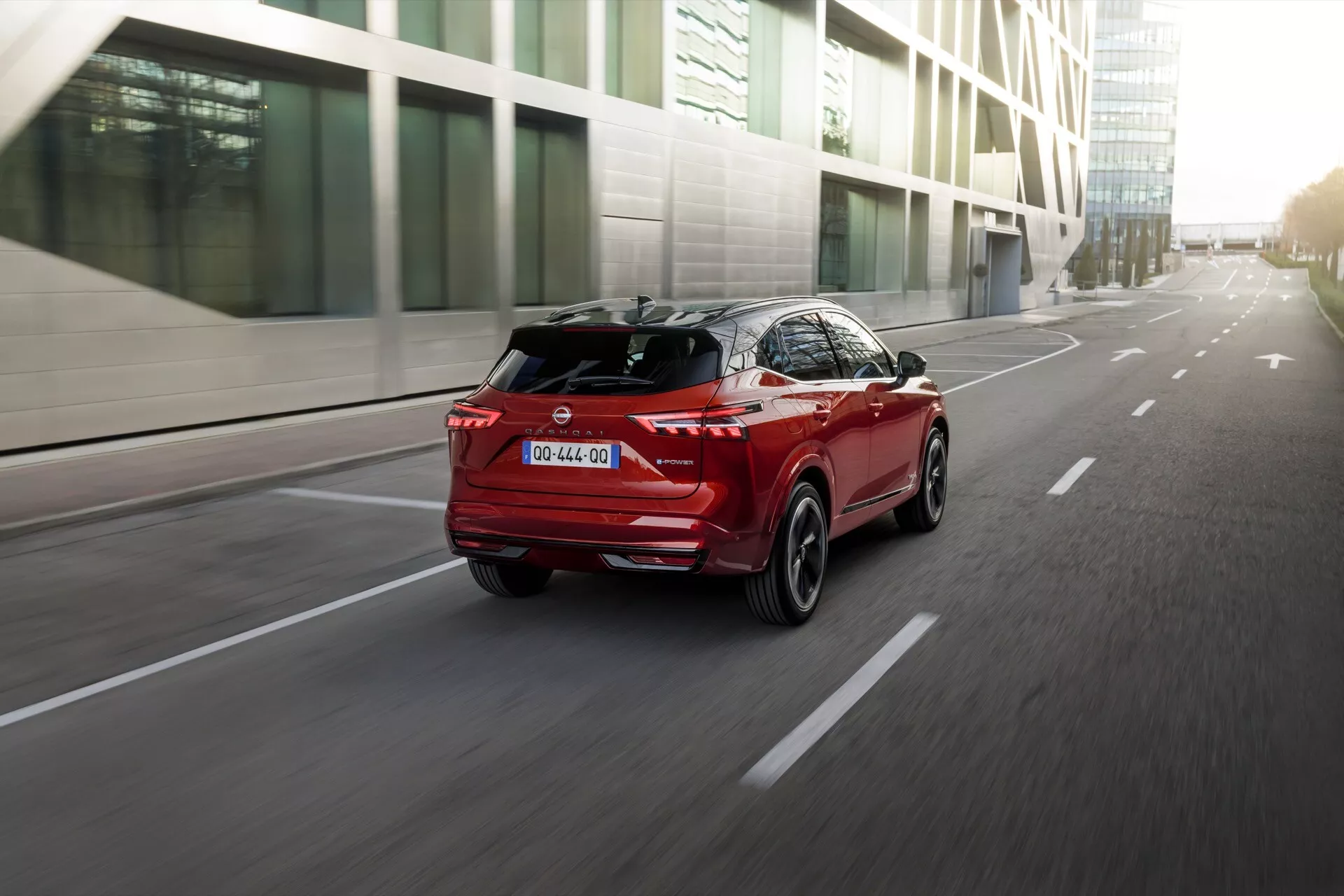 Foto dinâmico de três quartos traseiros de um SUV Nissan Qashqai J12-Facelift 2024 vermelho, viajando por uma estrada asfaltada, com faróis traseiros angulares bem iluminados, um spoiler traseiro proeminente e um difusor traseiro. O ângulo ligeiramente baixo acentua a altura e a postura do carro, com uma antena de barbatana de tubarão visível no teto e acabamento cromado nas partes inferiores das portas. O fundo urbano moderno está desfocado, destacando o caráter esportivo e as linhas de design avançadas do SUV.
