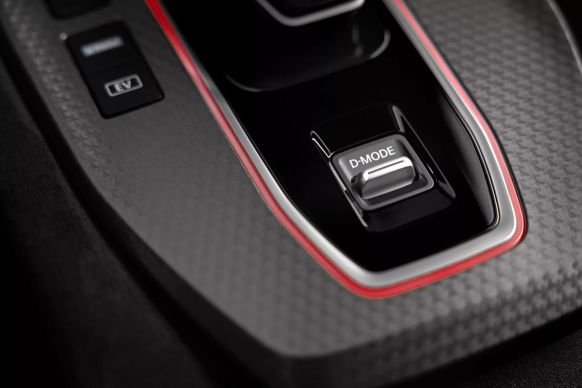 Vista aproximada da consola central do interior do Nissan Qashqai 2024 (J12-facelift), mostrando um seletor de marchas rotulado como 'D-MODE' inserido em um painel com textura de fibra de carbono, acabamento brilhante e destaque vermelho. O design sugere desempenho esportivo. A consola também apresenta dois botões, um indicando estabilidade ou dinâmica do veículo e outro possivelmente para ligar/desligar o motor.