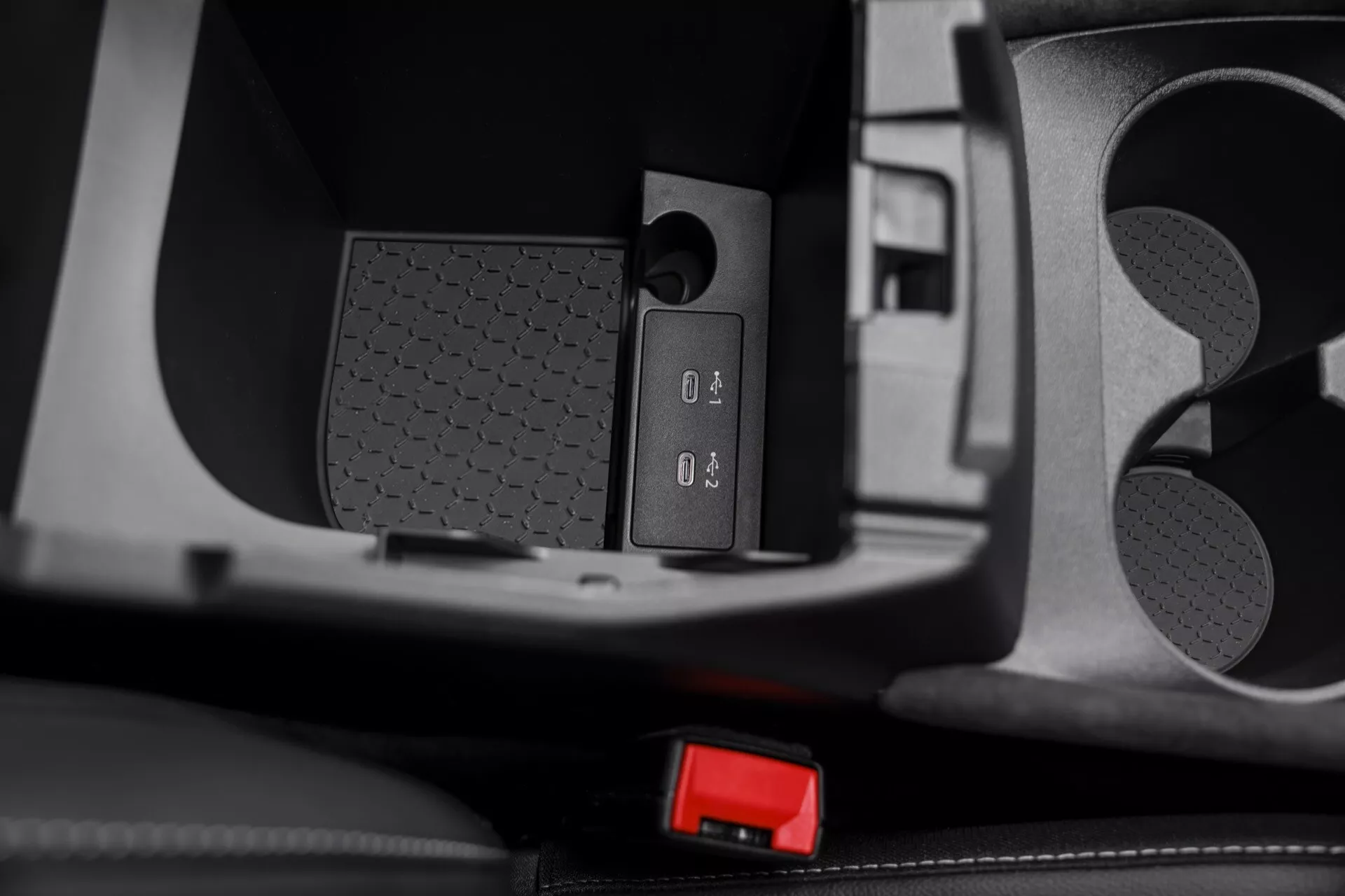 Vista interna de close-up da área da consola central do Nissan Qashqai 2024 (J12-facelift). A imagem mostra controles detalhados com sinais de mais (+) e menos (-), provavelmente para controle de volume do sistema de entretenimento. Ao lado dos controles há uma superfície texturizada, possivelmente um elemento de aderência ou design. A imagem também inclui um fecho de cinto de segurança visível com um botão de pressão vermelho. A foto captura várias texturas e materiais, enfatizando os acabamentos contrastantes da consola e áreas circundantes, fazendo um estudo no design interior automotivo, controles do sistema de entretenimento e design do cinto de segurança.