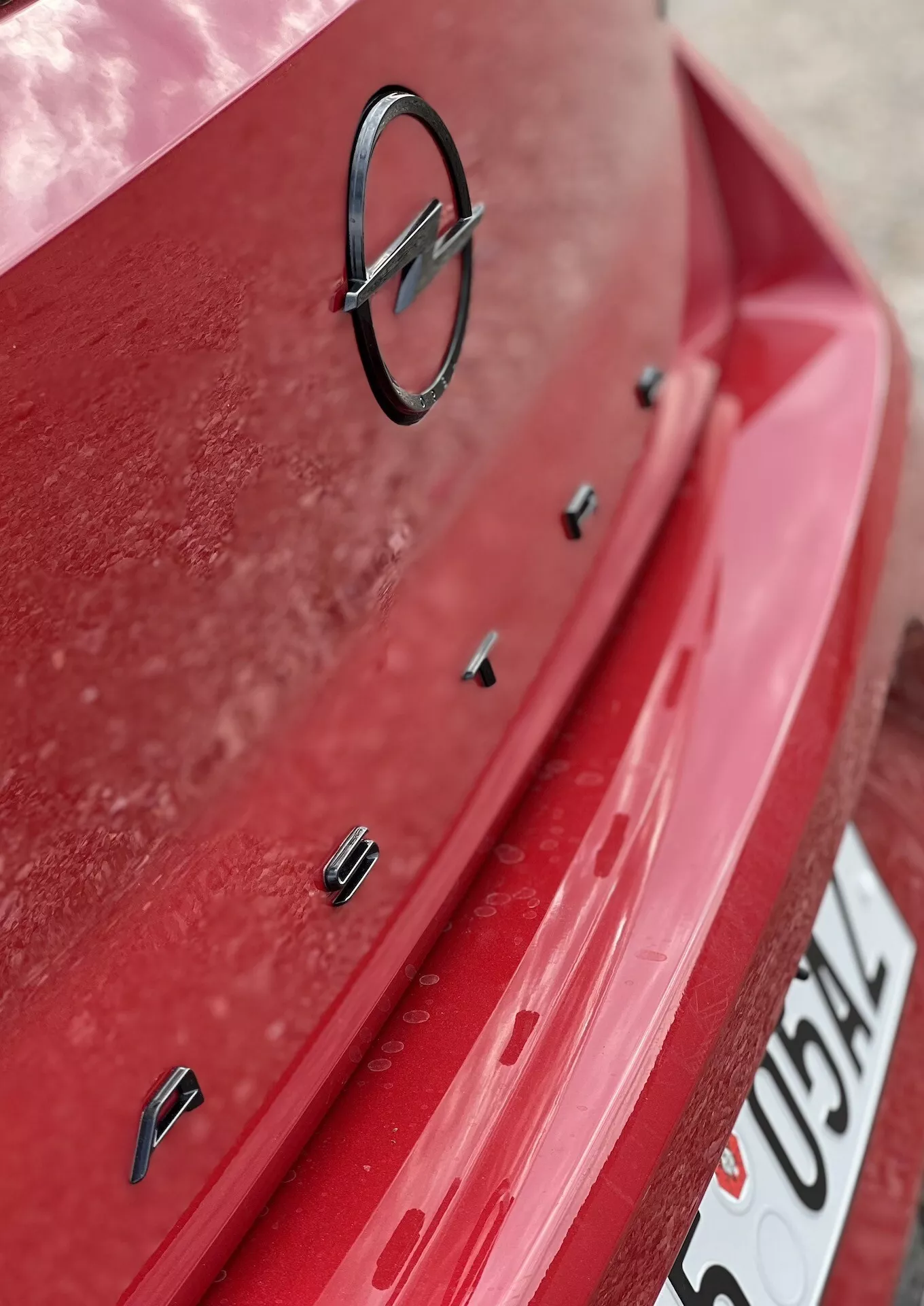 Vista de perto da seção traseira do Opel Astra 2022 com pintura vermelha e camada de poeira visível, mostrando o emblema do fabricante e sensores de assistência ao estacionamento