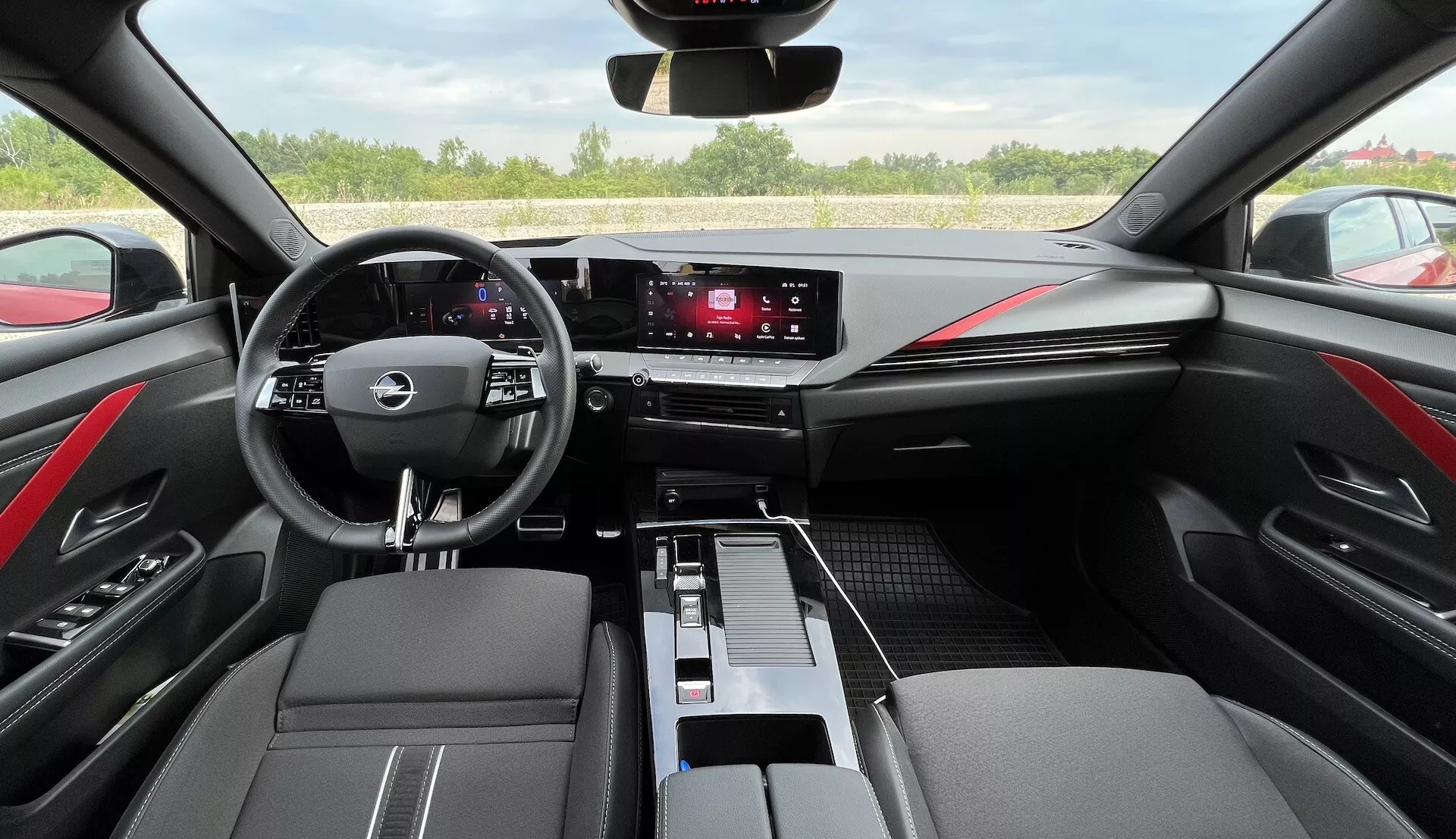Interior do Opel Astra 2022 exibindo volante multifunções com três raios, cluster de instrumentos digital, sistema de infotainment touchscreen, controles climáticos elegantes, acabamentos em duas tonalidades de preto com detalhes costurados em vermelho, e design moderno da cabine sob a perspectiva do condutor