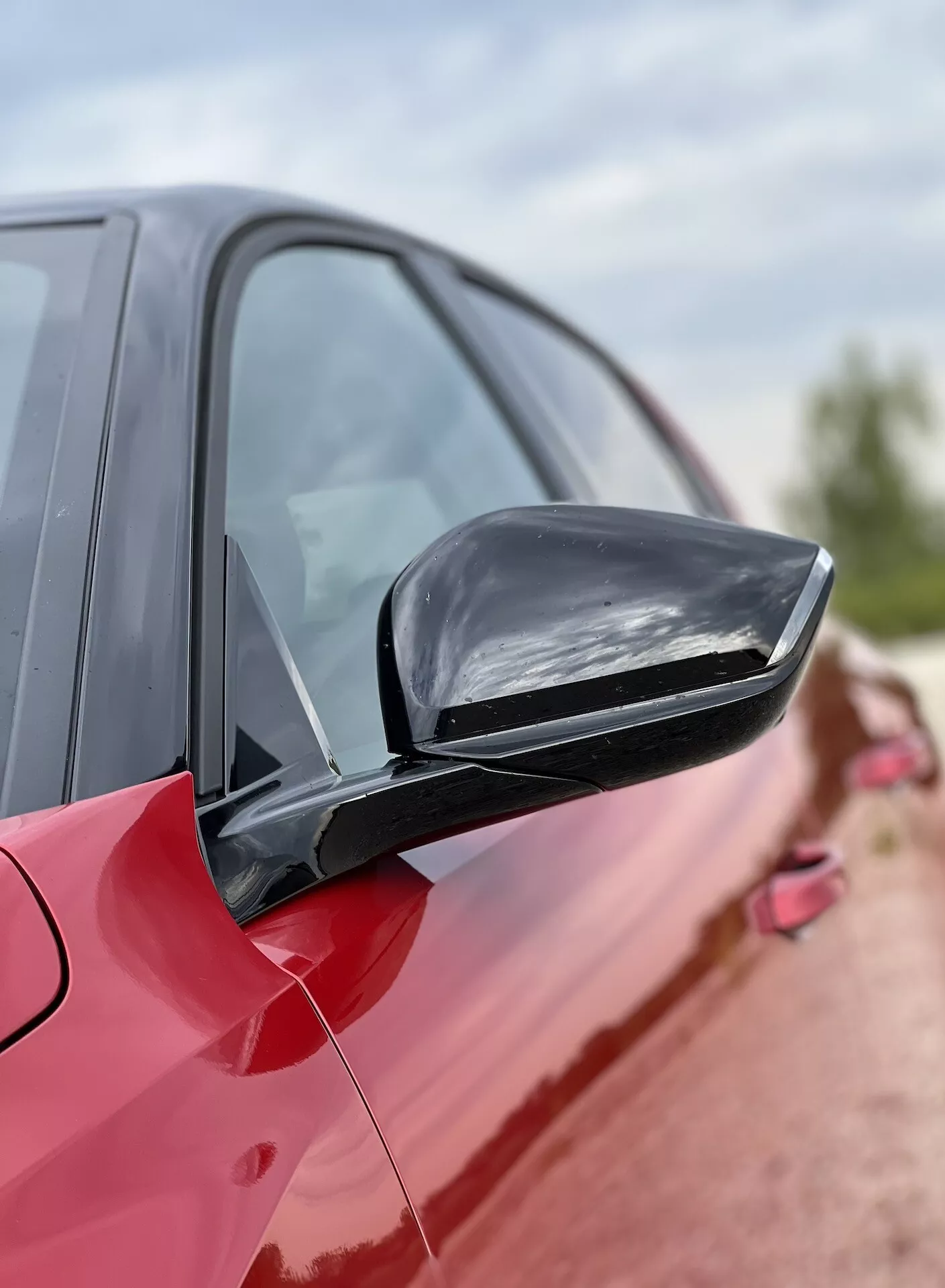 Close-up de um espelho lateral em um Opel Astra 2022 vermelho, destacando o design elegante e combinando com a cor e recursos tecnológicos potenciais contra uma silhueta de carro desfocada