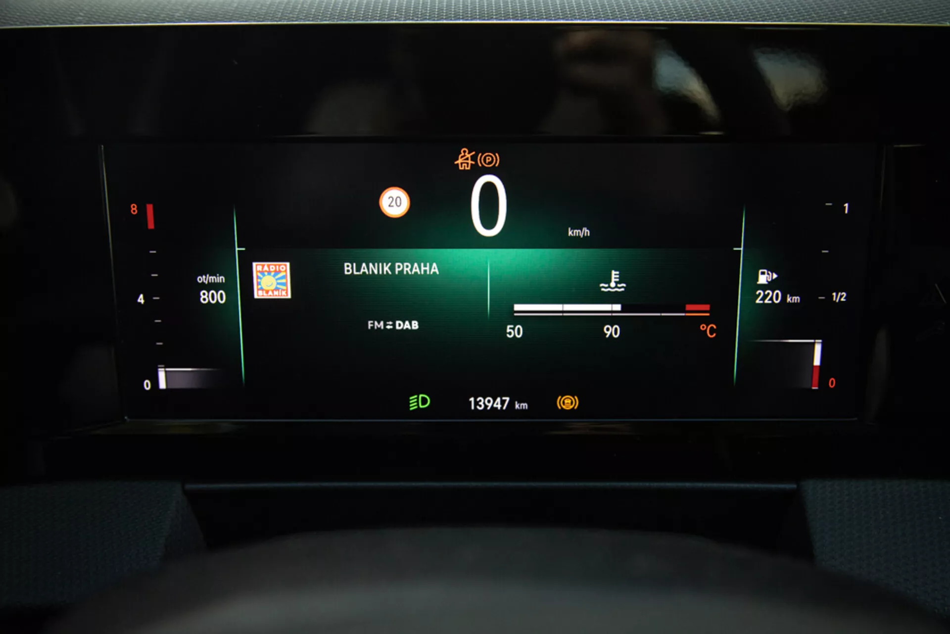 Vista do painel digital da Opel Astra 2022 destacando o cluster de instrumentos com RPM, nível de bateria, velocímetro, indicador de combustível, temperatura externa, estação de rádio 'BLANIK PRAHA', e leitura do odômetro a partir da perspectiva do assento do motorista.