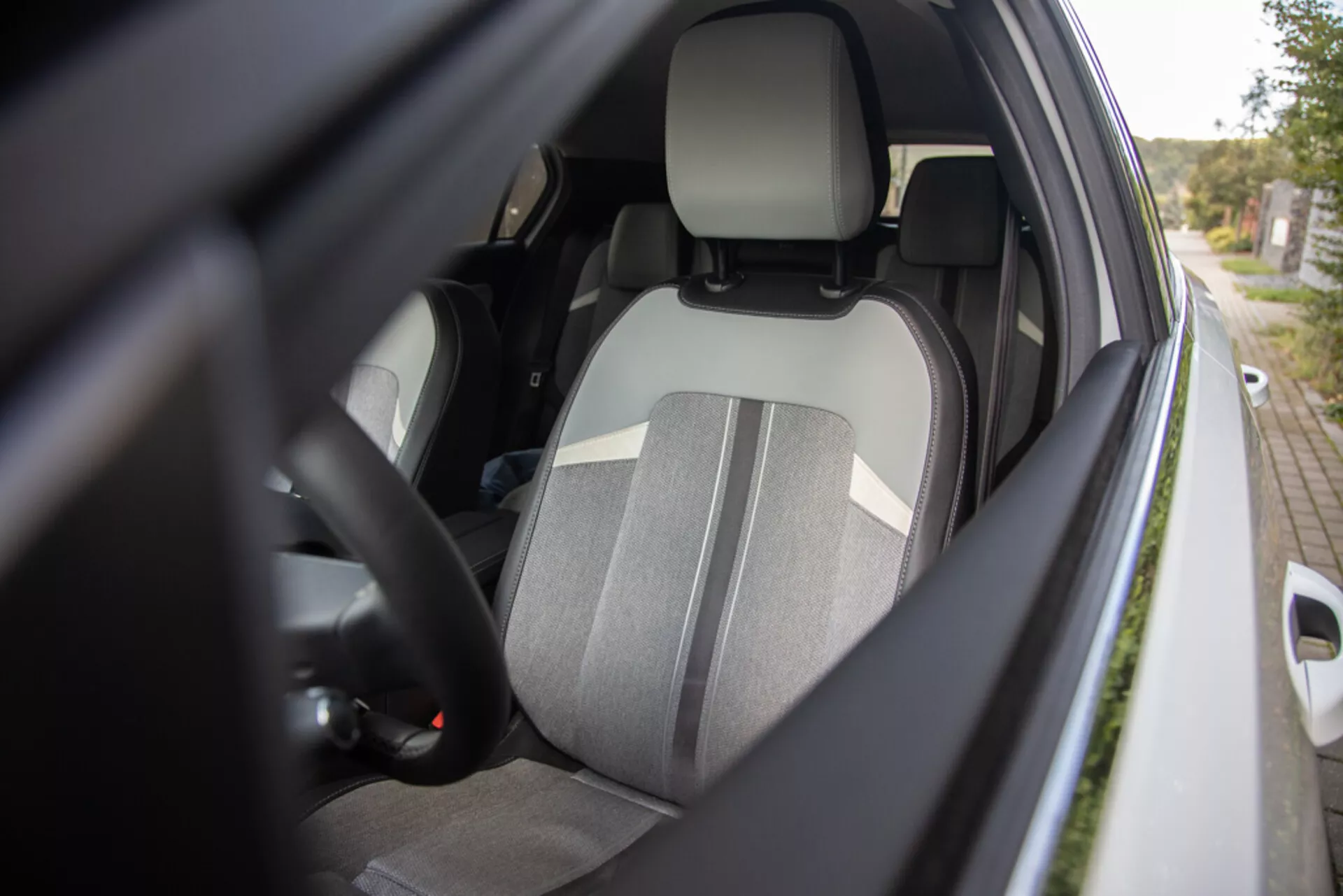 Vista detalhada do interior do lado do motorista do Opel Astra 2022 a partir de um ângulo lateral externo mostrando o assento do motorista em tecido de dois tons, encosto de cabeça integrado, parte do volante com controles, vislumbre do painel de instrumentos, cinto de segurança conectado, interior da porta com possíveis controles para janelas elétricas e a caixa do espelho lateral na cor da carroceria enfatizando ergonomia, segurança e recursos de conveniência.
