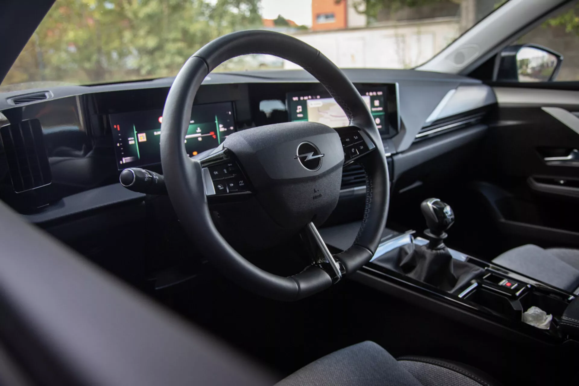 Interior do Opel Astra 2022 mostrando tecnologia moderna de painel digital, volante multifuncional, sistema de infotenimento de tela dupla larga e alavanca de câmbio manual para desempenho esportivo, visto do banco traseiro em direção à área do motorista com acabamentos premium